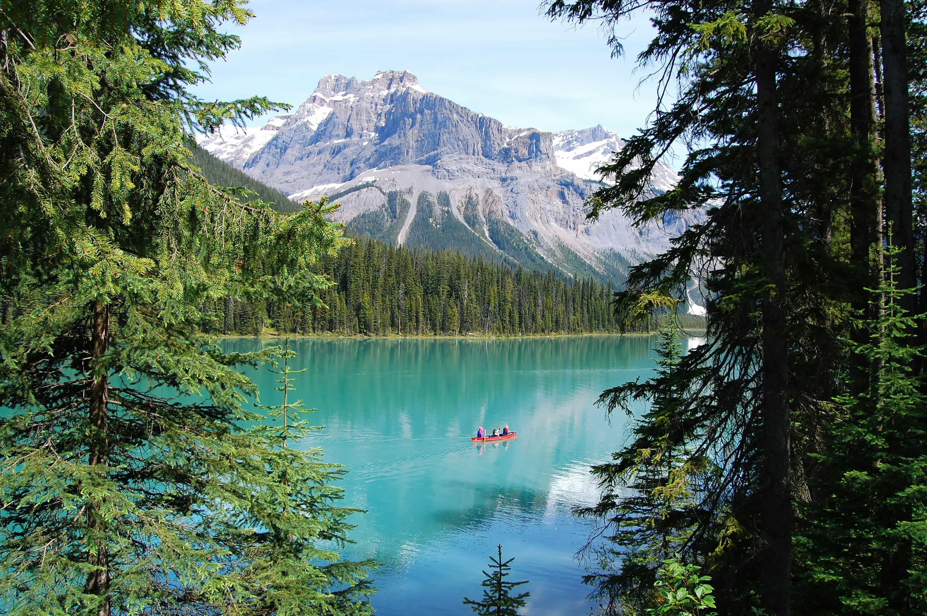 Turquoise lake surrounded by dense evergreen forest and snow-capped mountains, with a red canoe carrying people gliding across the calm water under a clear blue sky.
