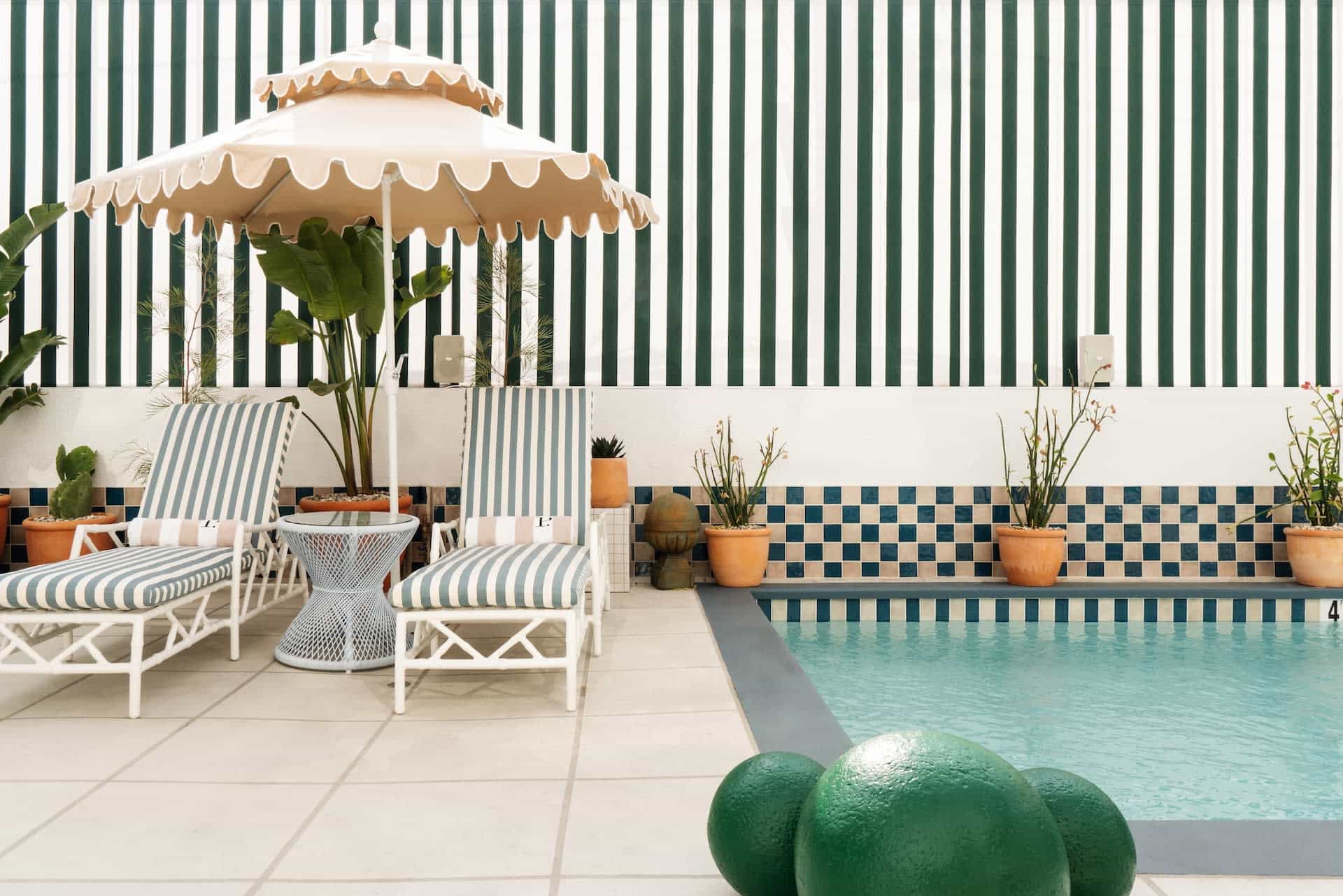 Chic poolside scene with striped lounge chairs, a scalloped umbrella, potted plants, and a turquoise pool framed by green-and-white striped walls and tiled accents.