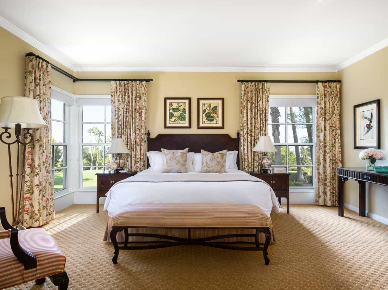 Elegant traditional bedroom with floral curtains, framed botanical art, and a large wooden bed dressed in white linens and gold-accented pillows overlooking a garden view.