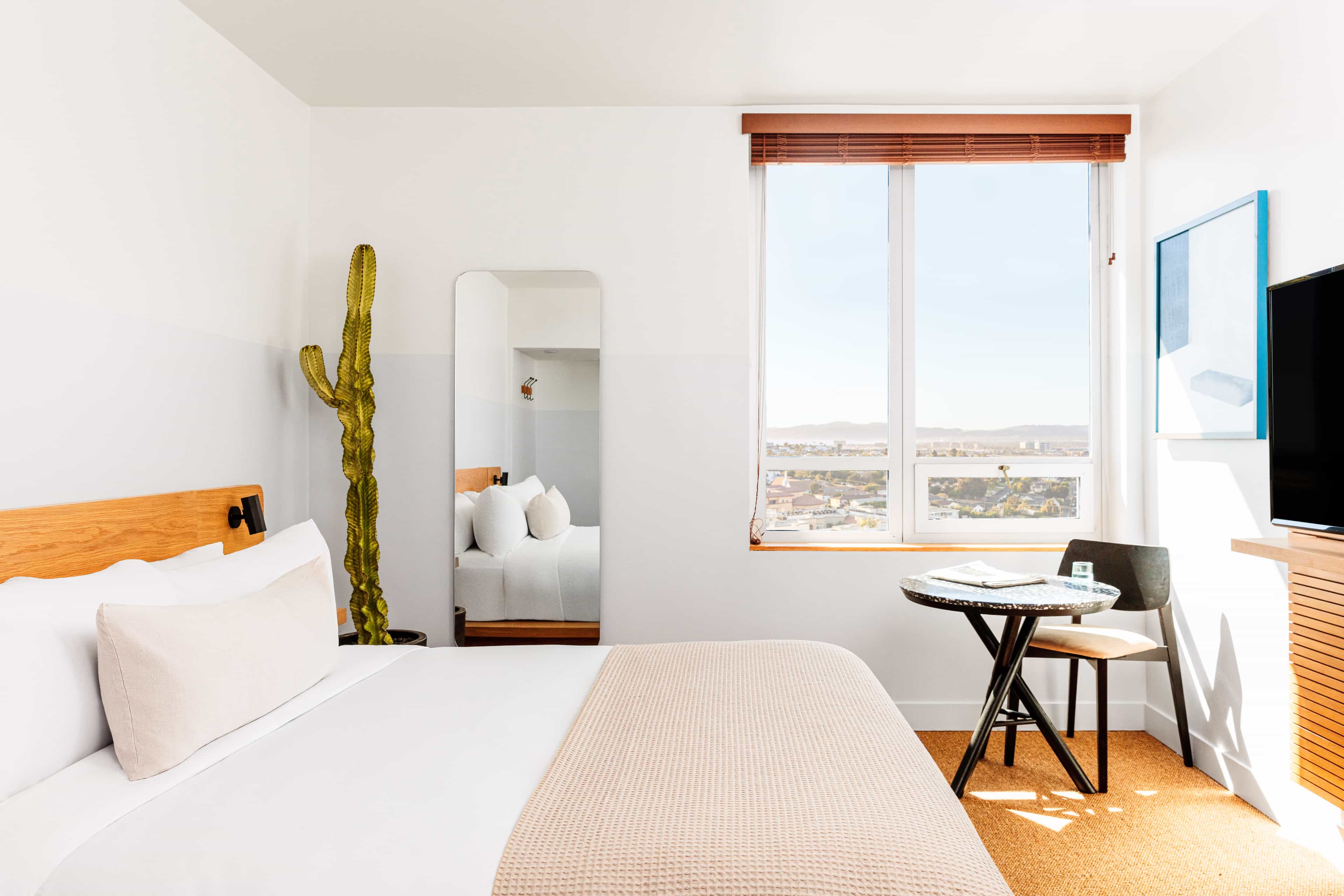 Minimalist hotel room with a cactus, light wood furnishings, and a large window overlooking the city under bright natural light.