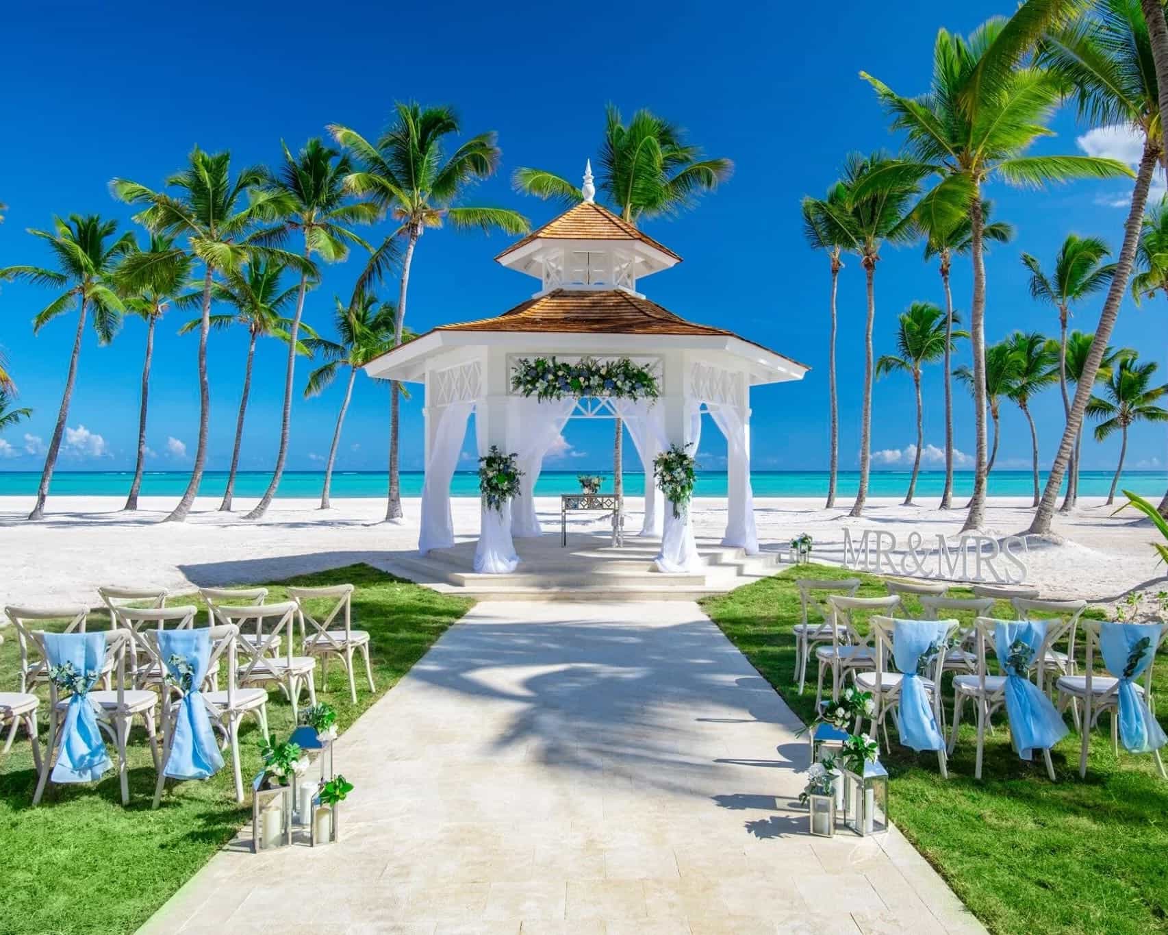 white gazebo on a white sand beach with chairs lining the walkway, and palm trees and ocean behind under blue skies during day