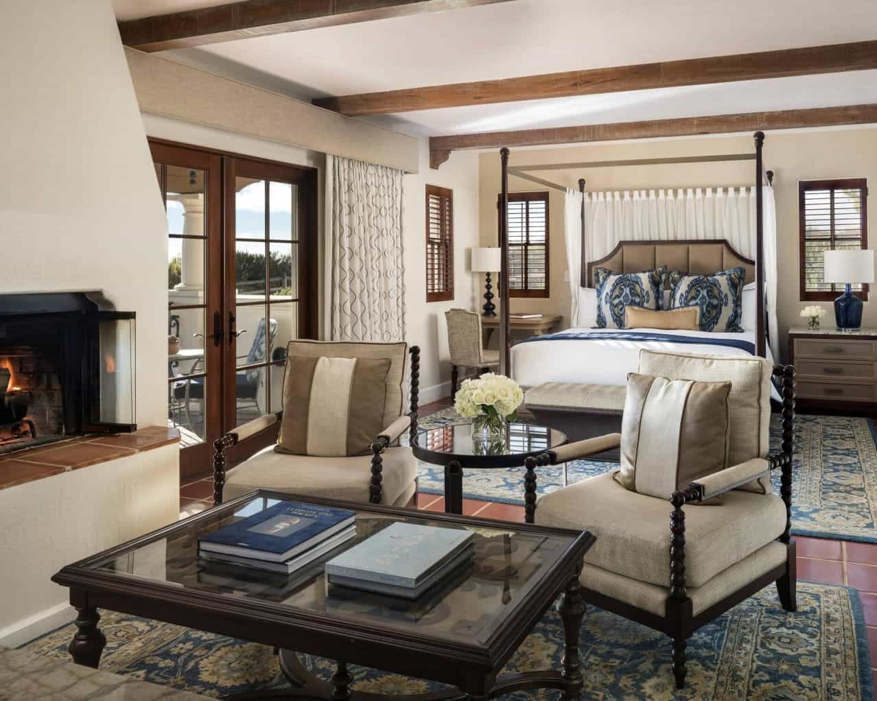 a contemporary hotel room with foud poster bed, decorative pillows, coffee table and chairs next to a fireplace, and wood beam ceiling