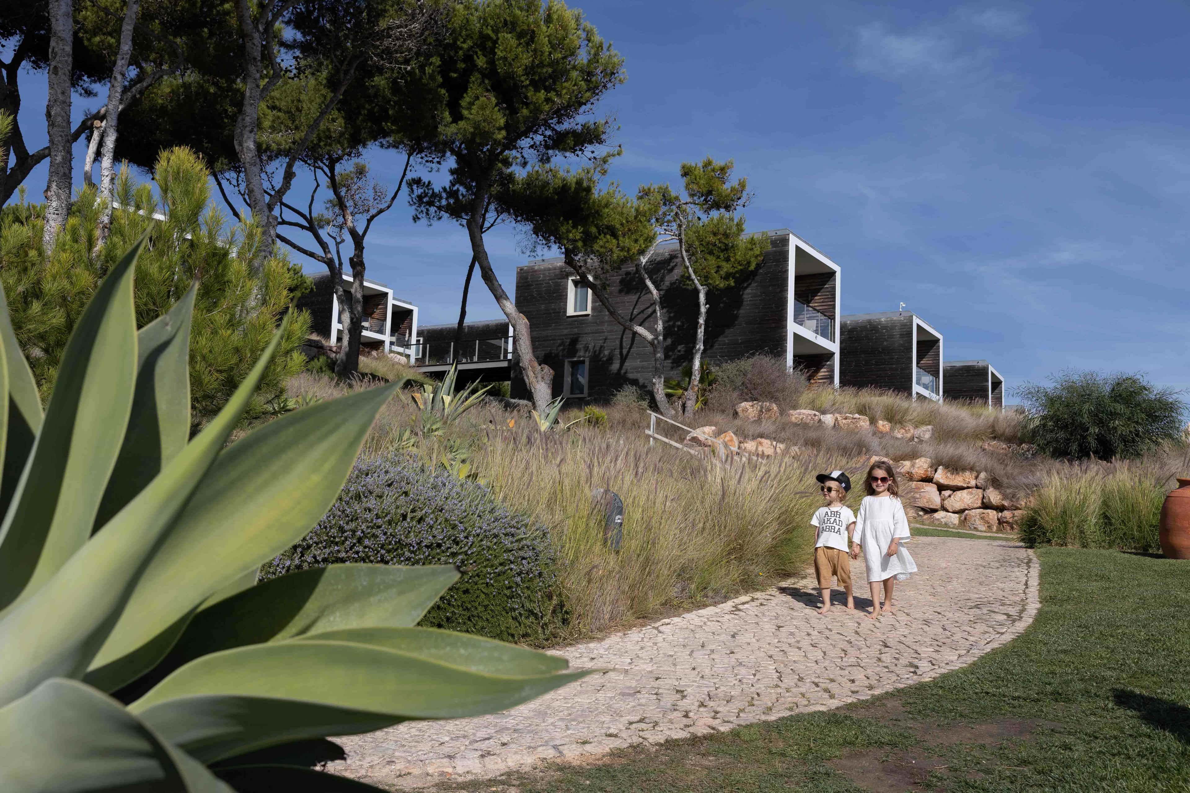 Two children walking on a stone path surrounded by greenery and modern villa-style buildings at a coastal resort with blue skies above.
