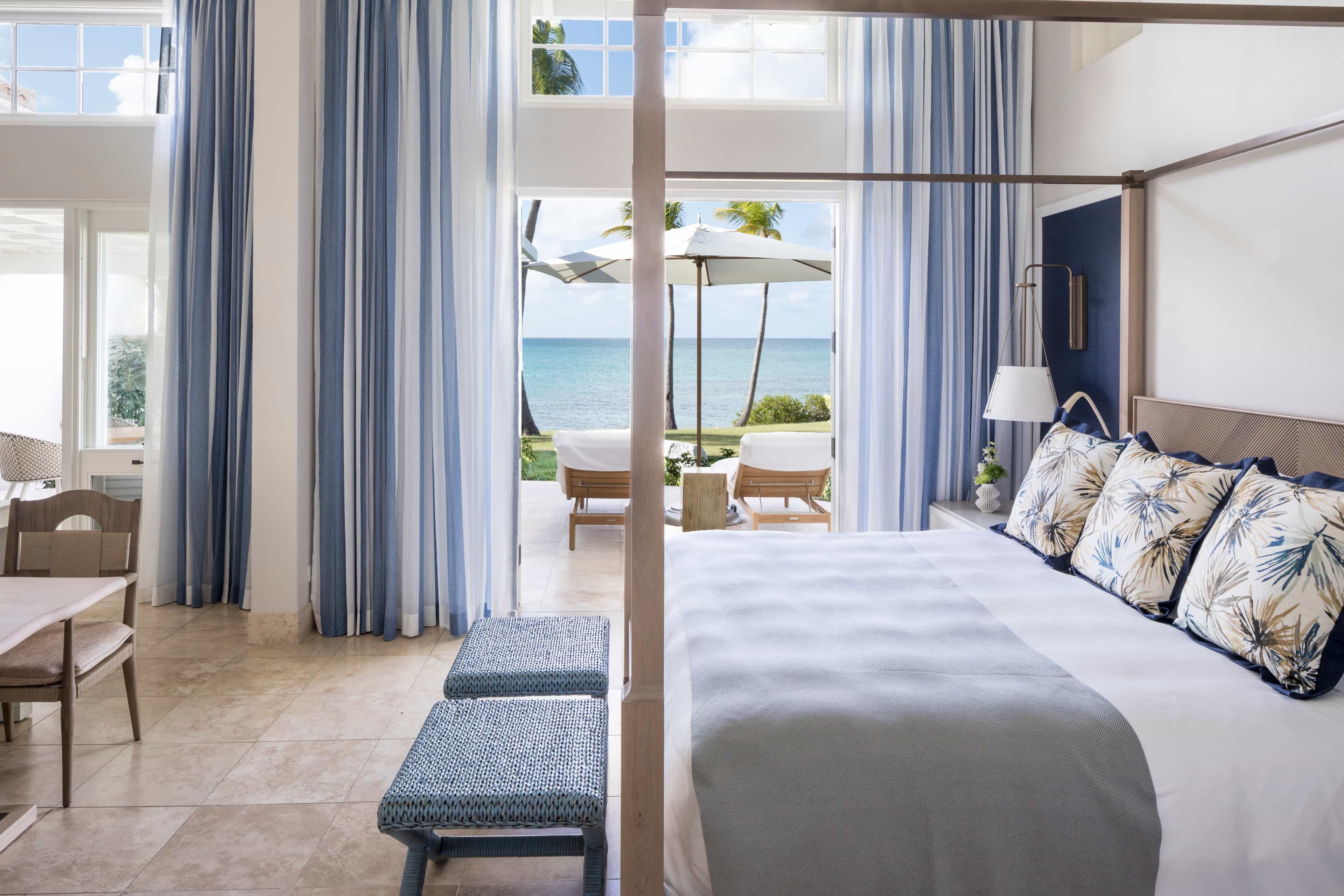 A bright, coastal-inspired bedroom with blue-and-white striped curtains, a four-poster bed, and patterned pillows, opening onto a terrace with sun loungers, an umbrella, and views of palm trees and the turquoise ocean.