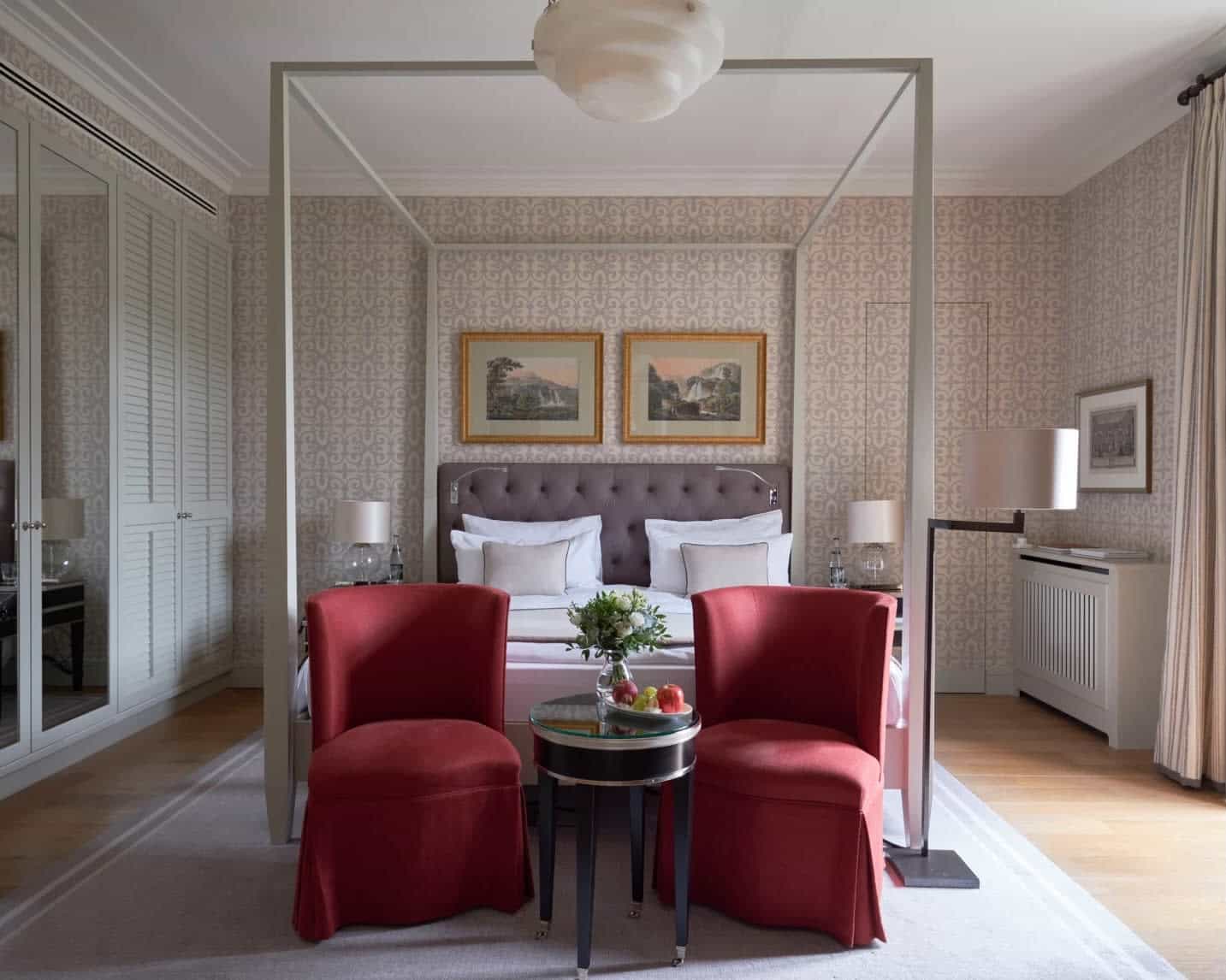 four poster bed with grey headboard and pictures above the bed, red chairs and side table at the foot of the bed, and floral wallpaper on the walls