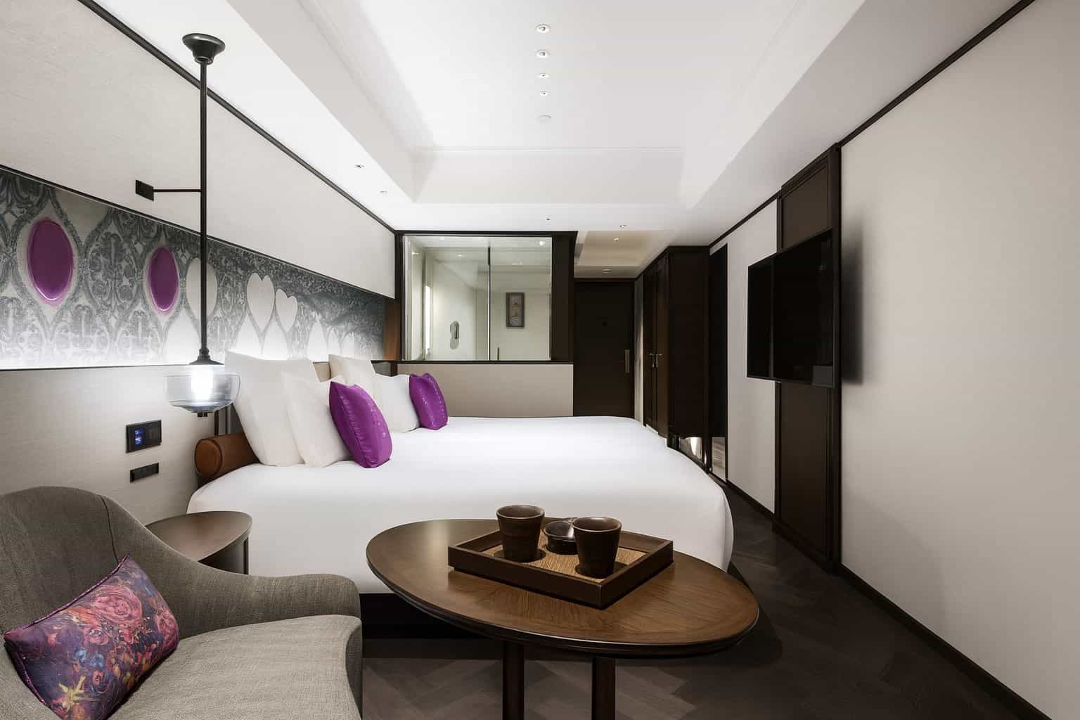 Contemporary hotel room with two white beds accented by purple pillows and a patterned headboard. A cozy seating area with a grey armchair and small wooden table sits in the foreground, while a large TV is mounted on the opposite wall.