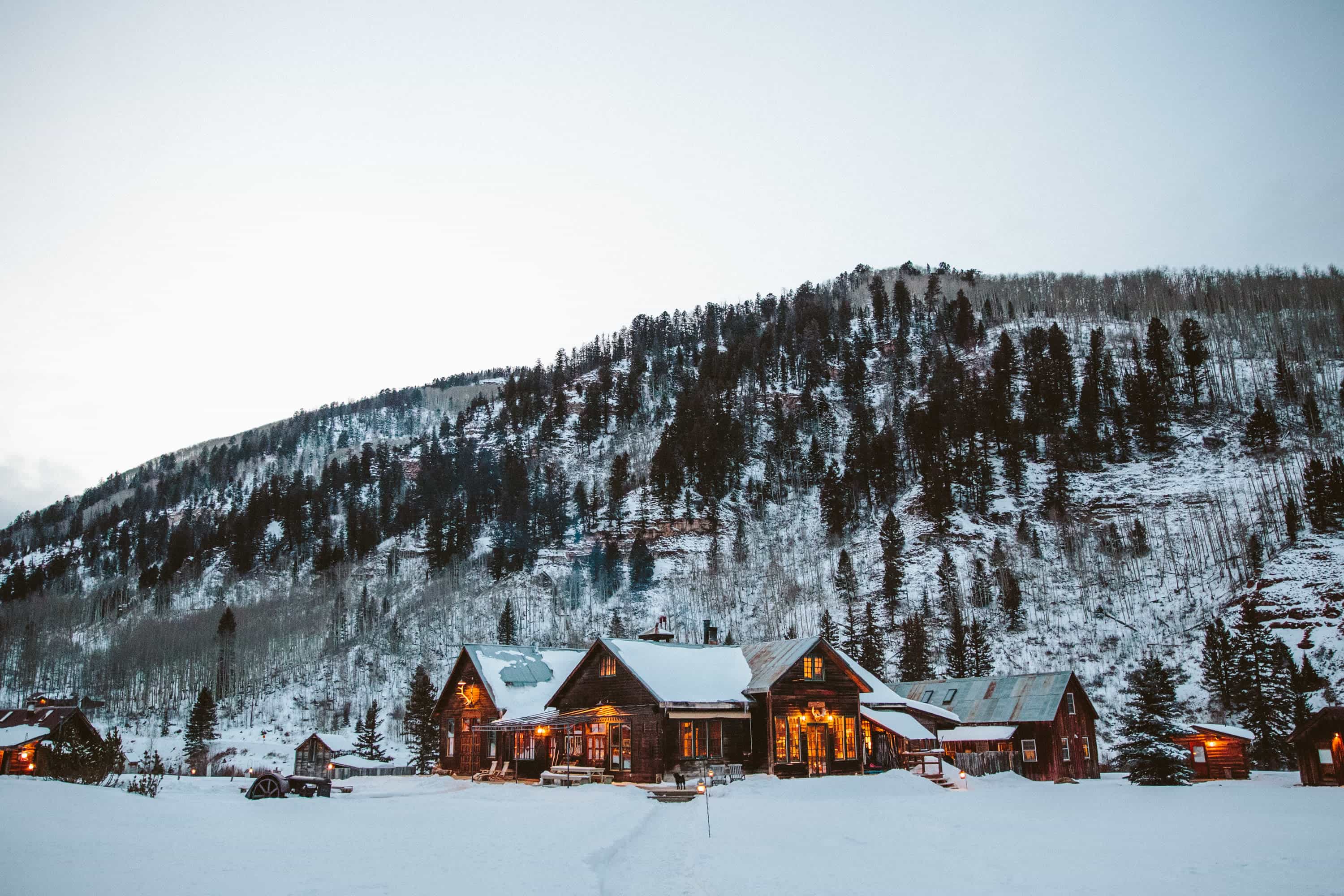 A cluster of rustic wooden cabins with warm glowing lights sits in a snowy valley at the base of a pine-covered mountain, creating a cozy winter lodge atmosphere.