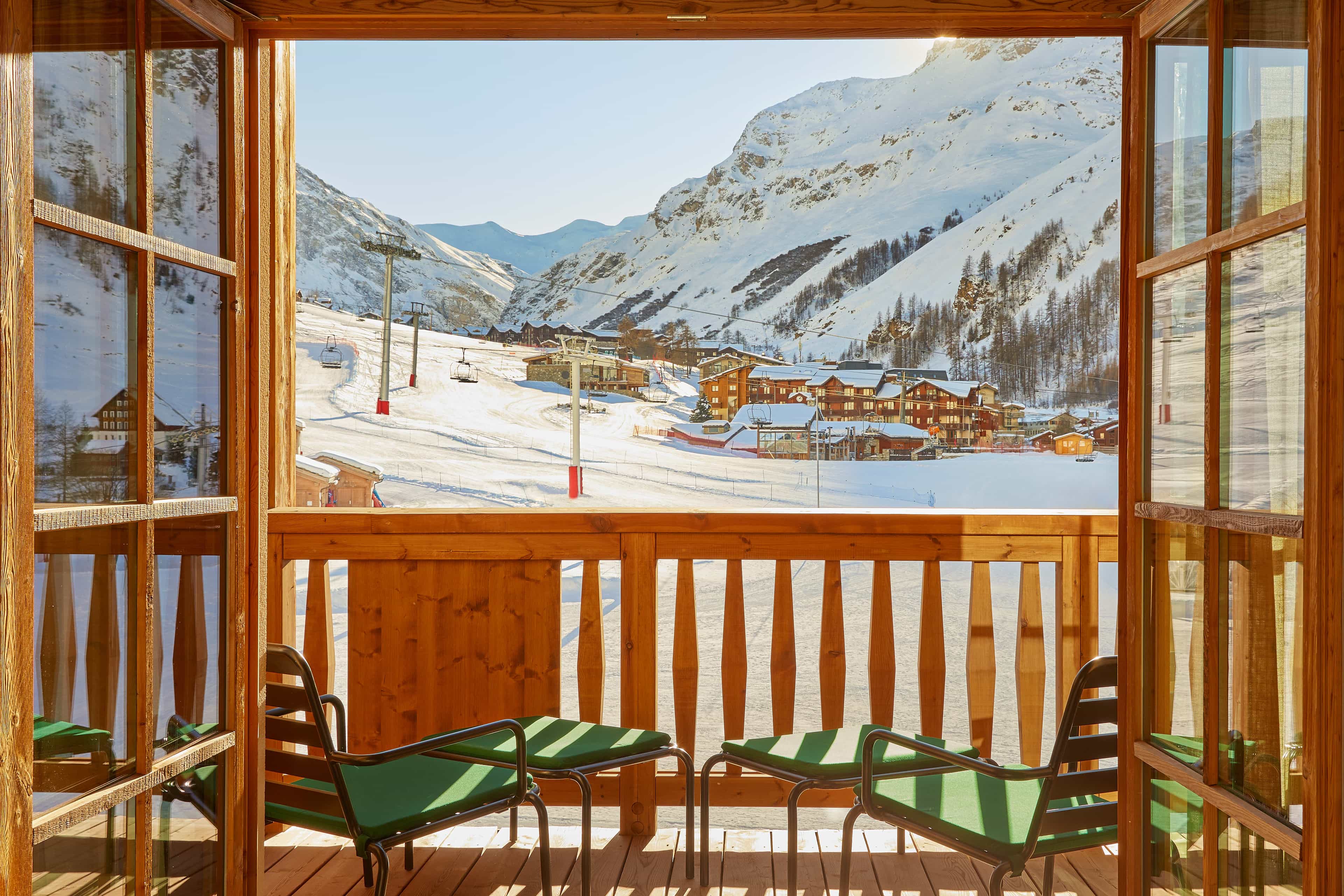 A wooden balcony with green chairs overlooking a snowy mountain ski resort, with ski lifts, chalets, and sunlit slopes framed by open wooden doors.