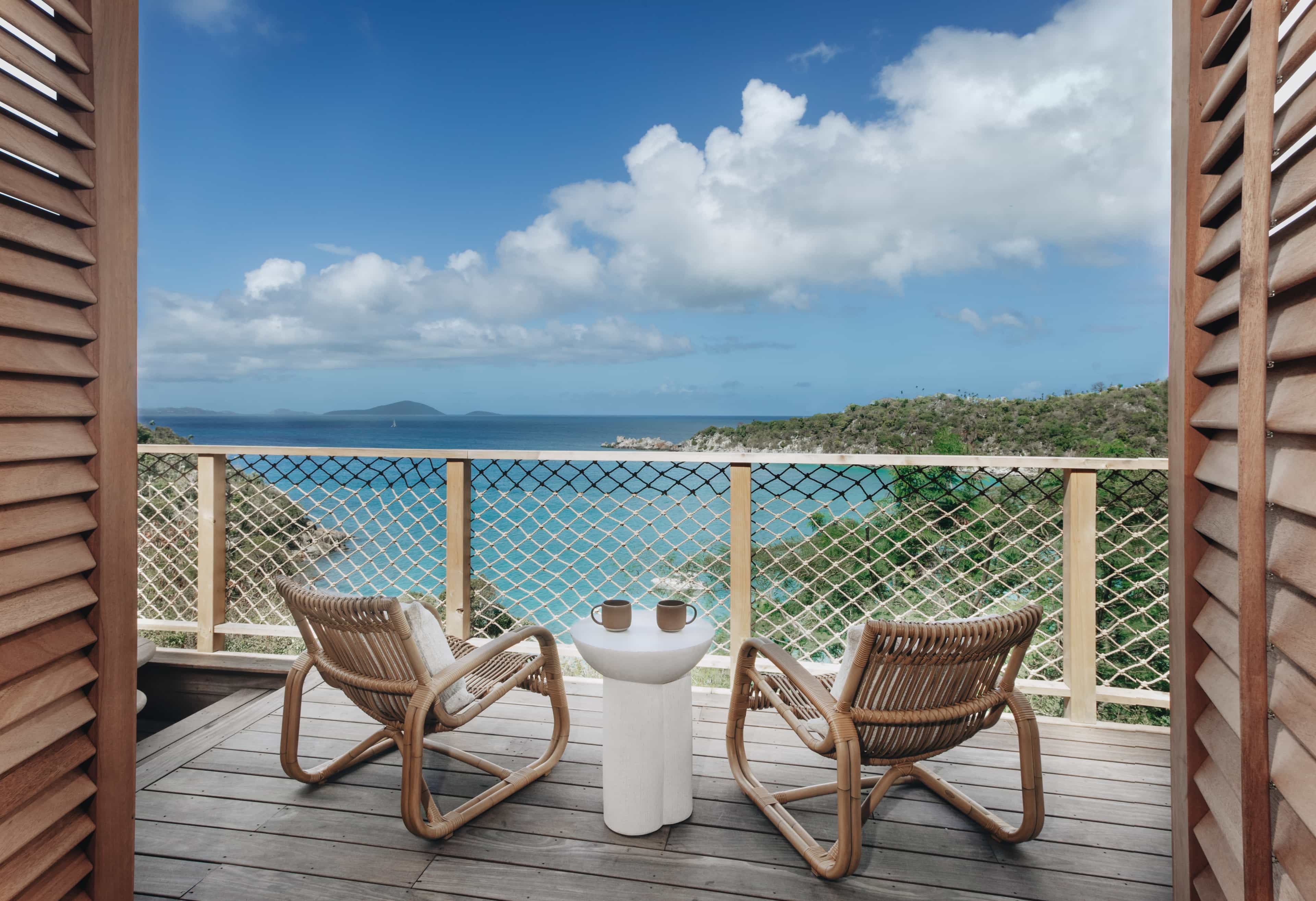 Two woven lounge chairs and a small table with mugs on a wooden deck overlooking turquoise water, green hills, and a blue sky with scattered clouds.