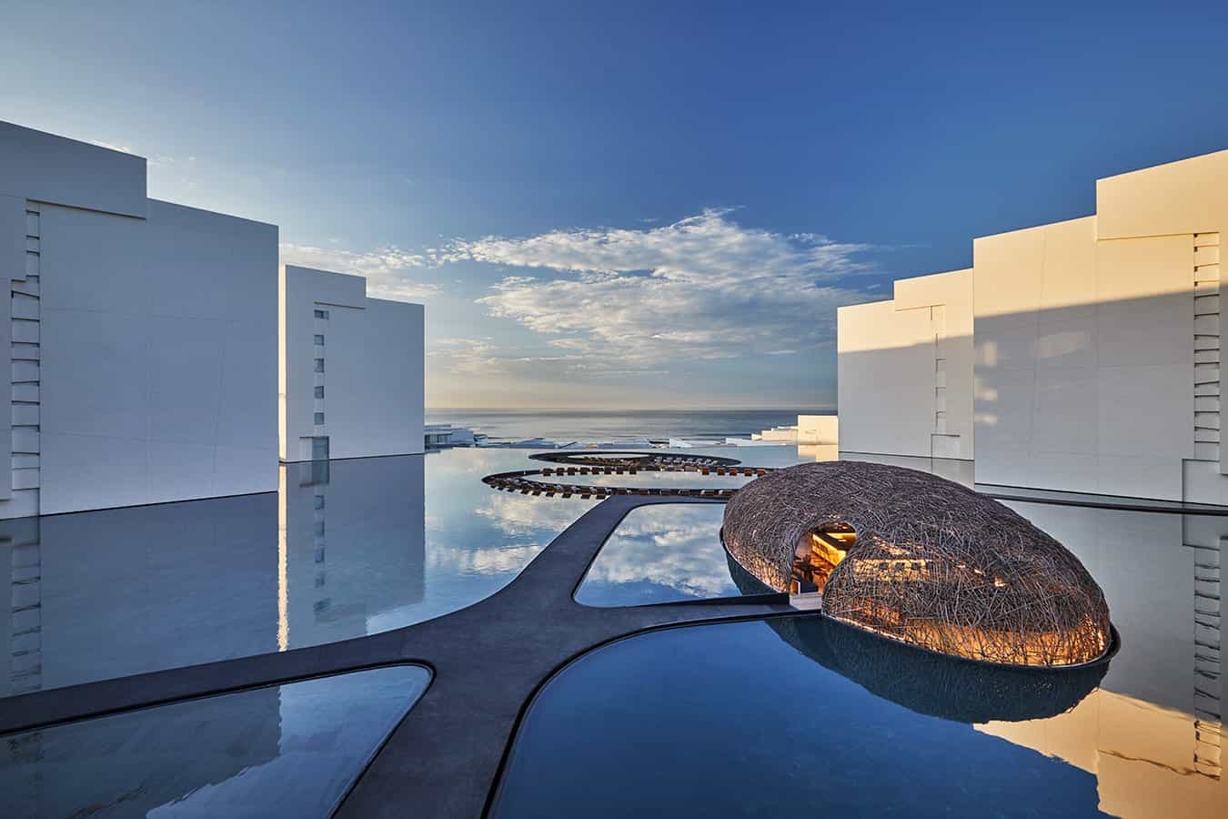 Striking modern courtyard with mirror-like reflecting pools, sculptural woven pavilion glowing at dusk, and crisp white geometric buildings framing open views toward the sea under a soft evening sky.