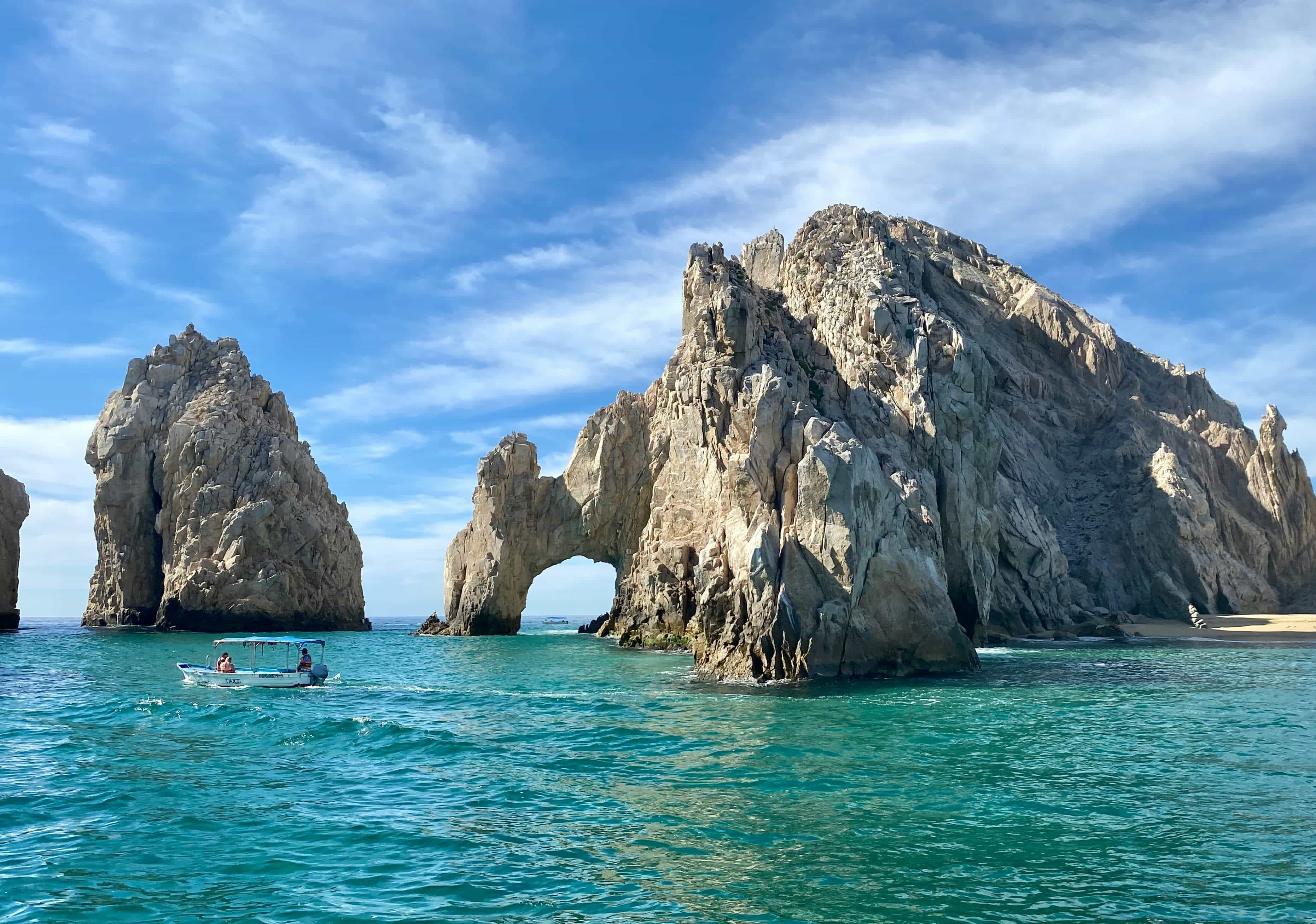 El Arco rock formation in Cabo San Lucas, with dramatic granite arches rising from turquoise water, a small boat passing nearby, and a bright blue sky with wispy clouds overhead.