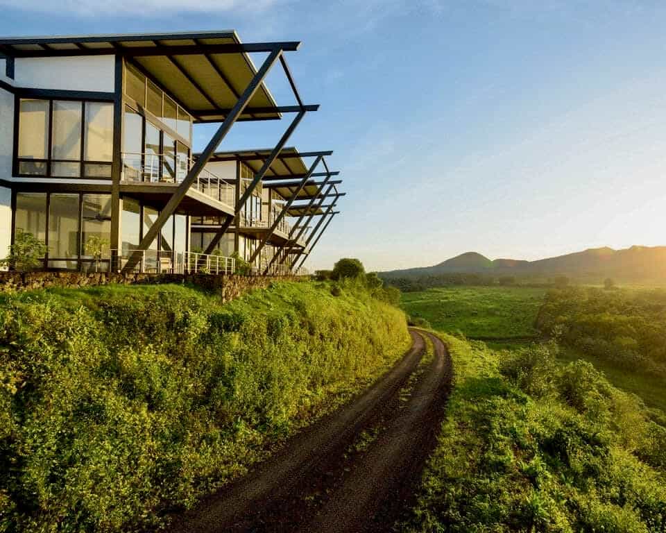 sun rishing over distant green mountains, with a modern lodge and dirt trail in the foreground