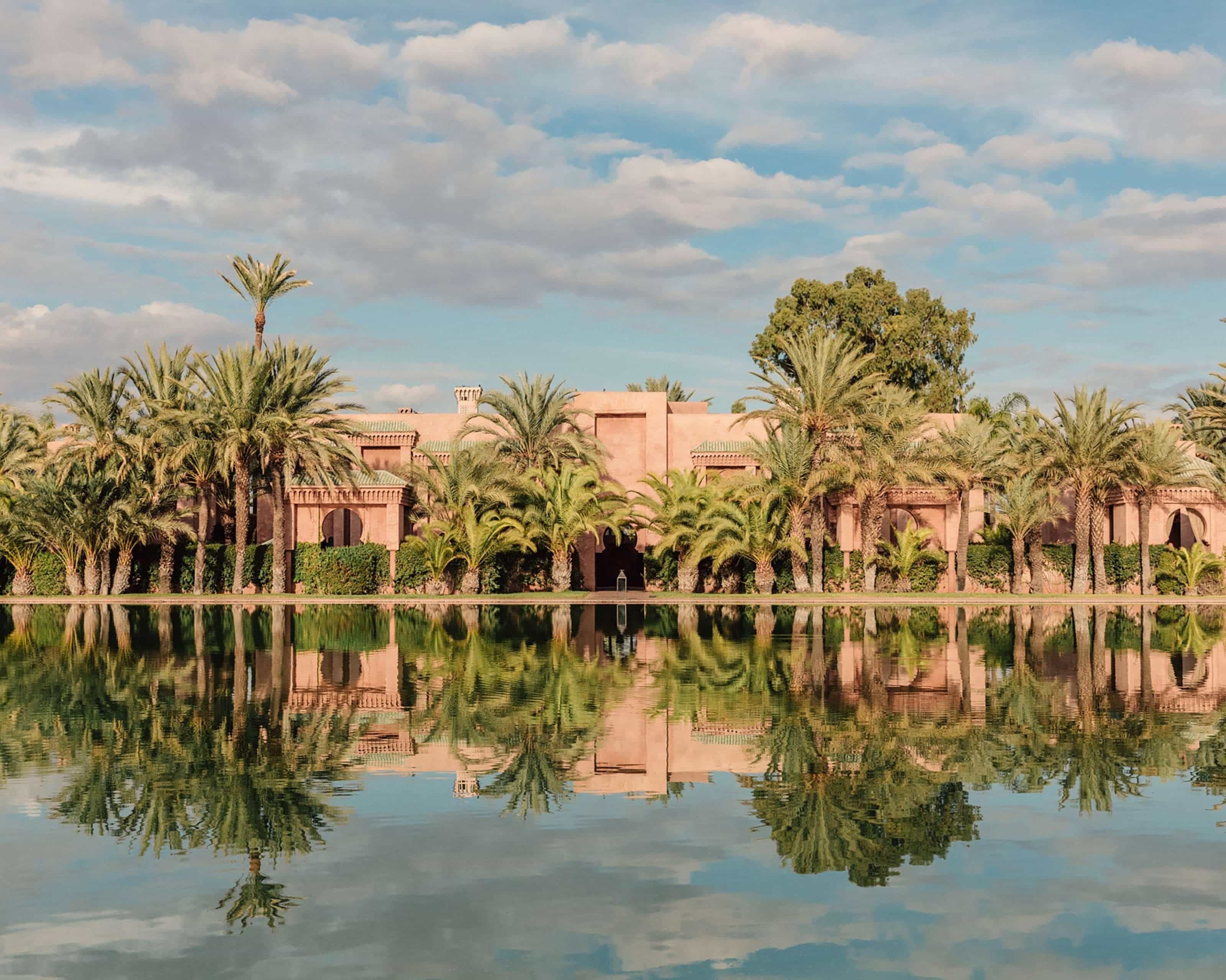 water reflecting a tan stucco building with palm trees in front and blue skies above