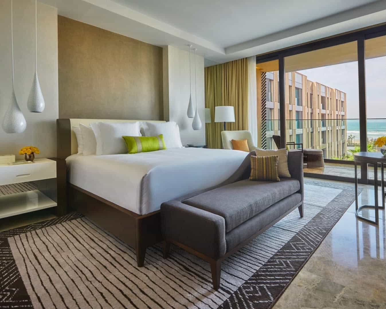 modern hotel room with white bed linens, grey bench, black striped rug, and ocean view out the windows