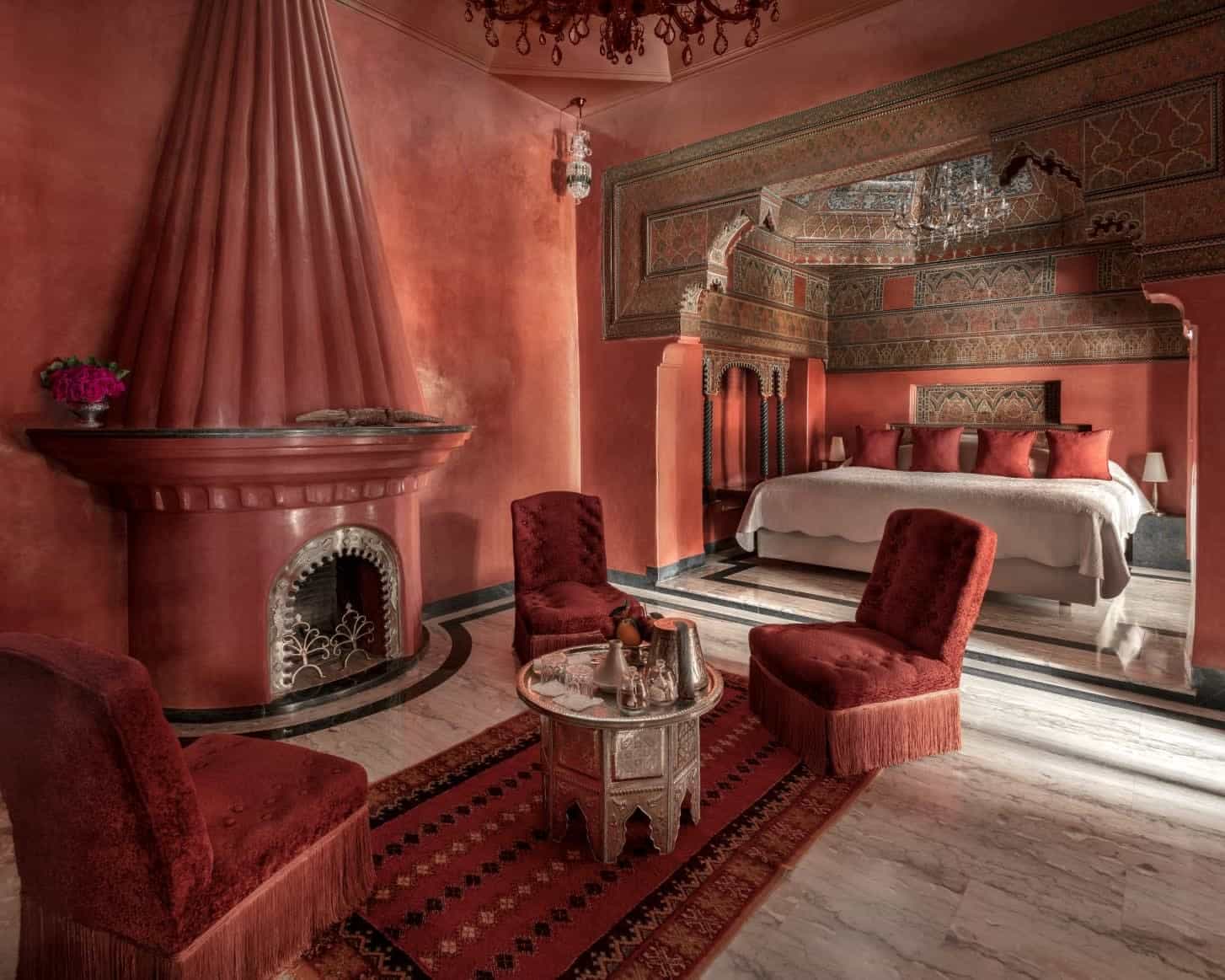ornate red hotel room with fireplace, chairs, and arched entryway to a white linen bed