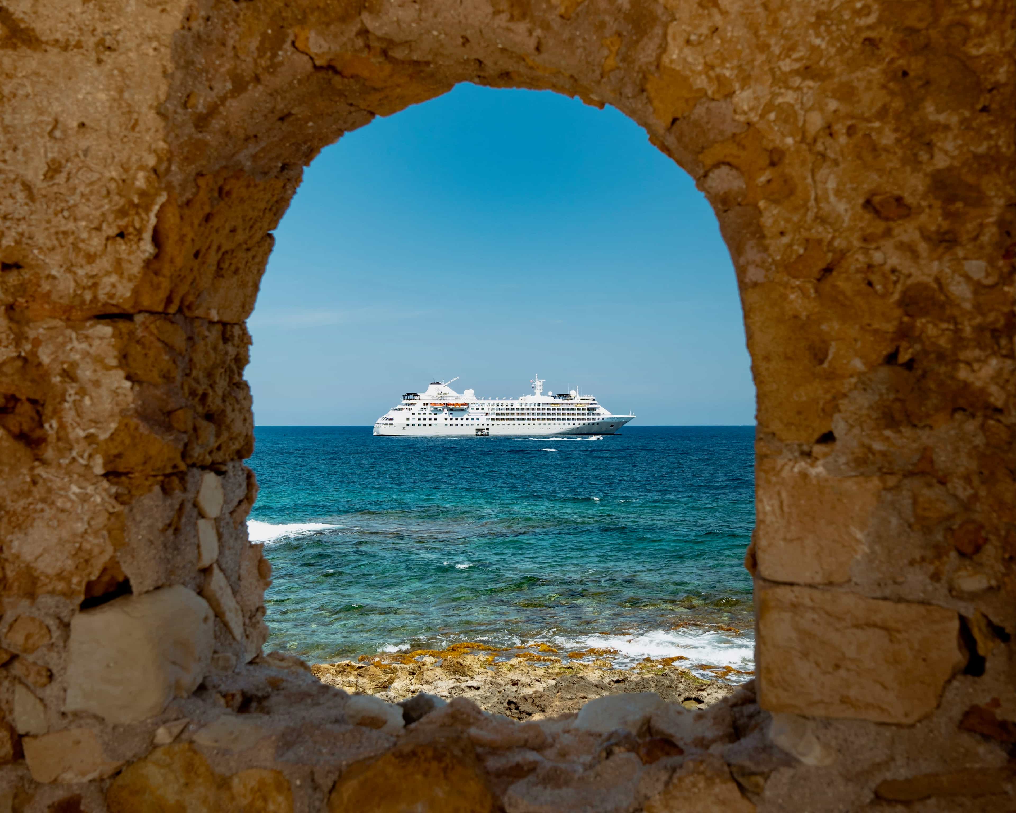 A cruise ship offshore in turquoise waters seen through an arched window in an ancient stone fort