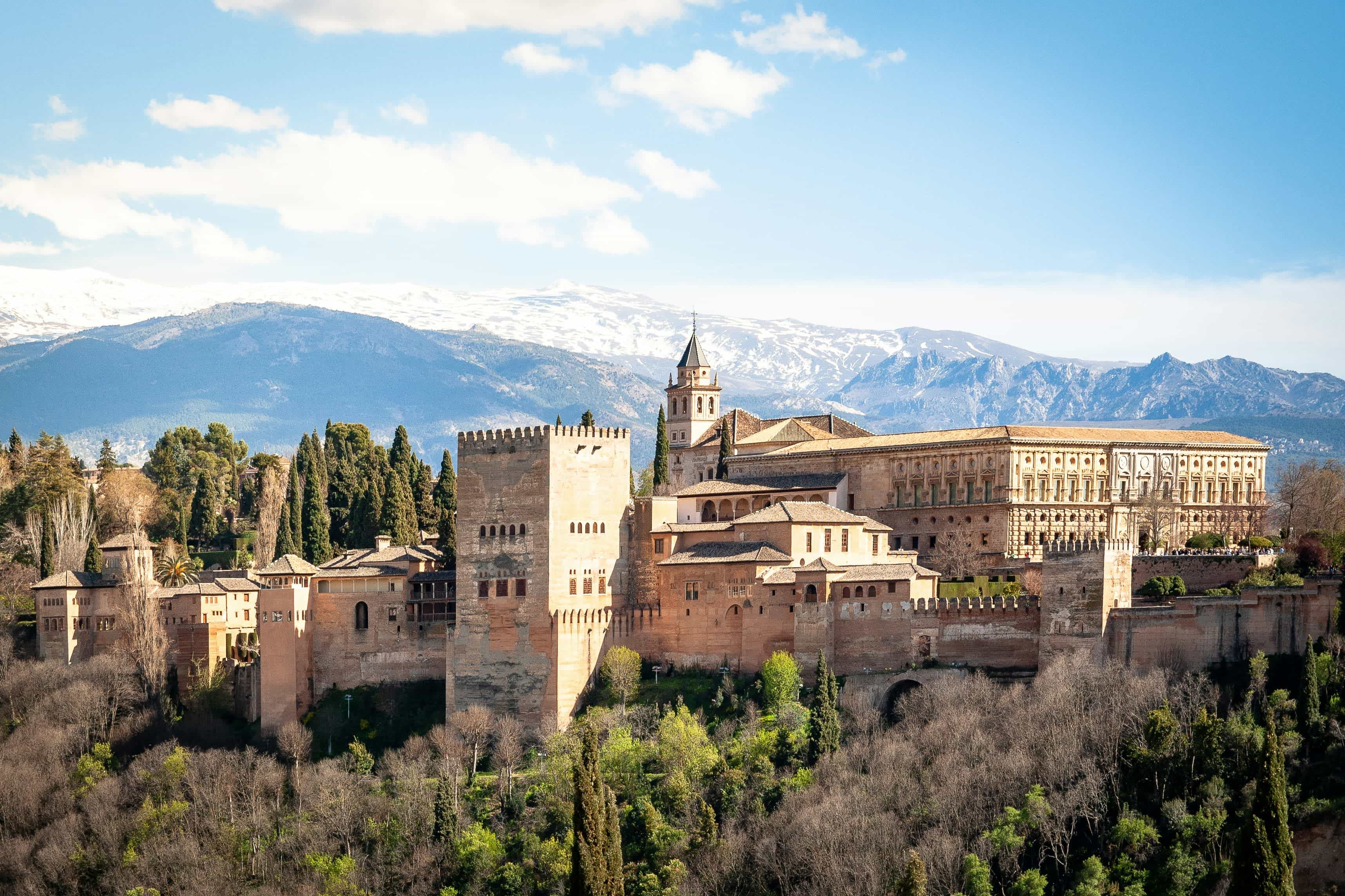 a cream colored fortress with trees in the foreground and snow capped mountains in the distance