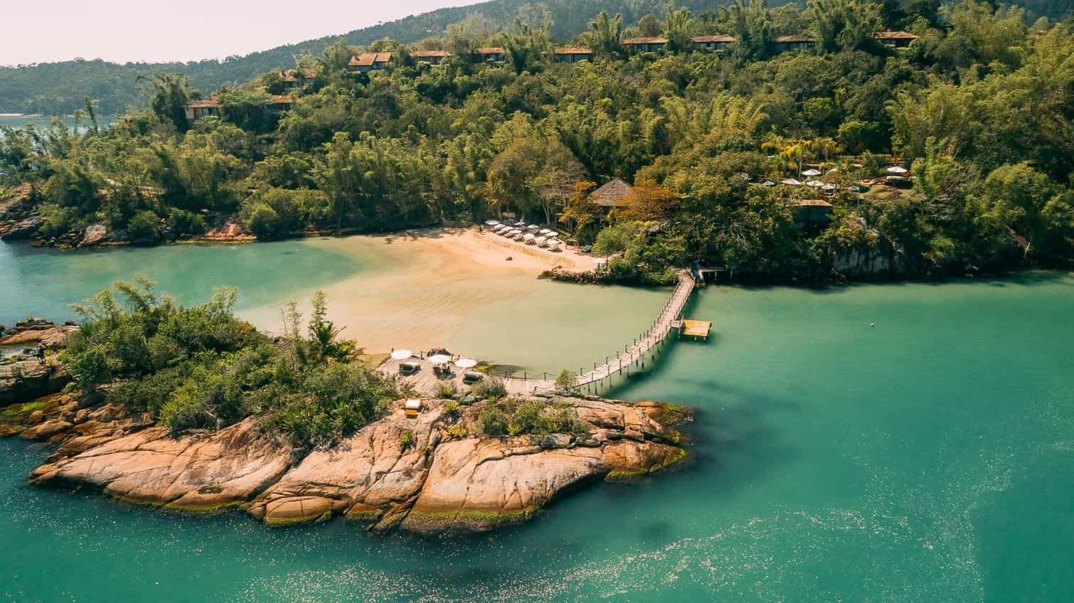 Aerial view of a secluded tropical beach with a wooden pier, turquoise water, and jungle-covered hills.