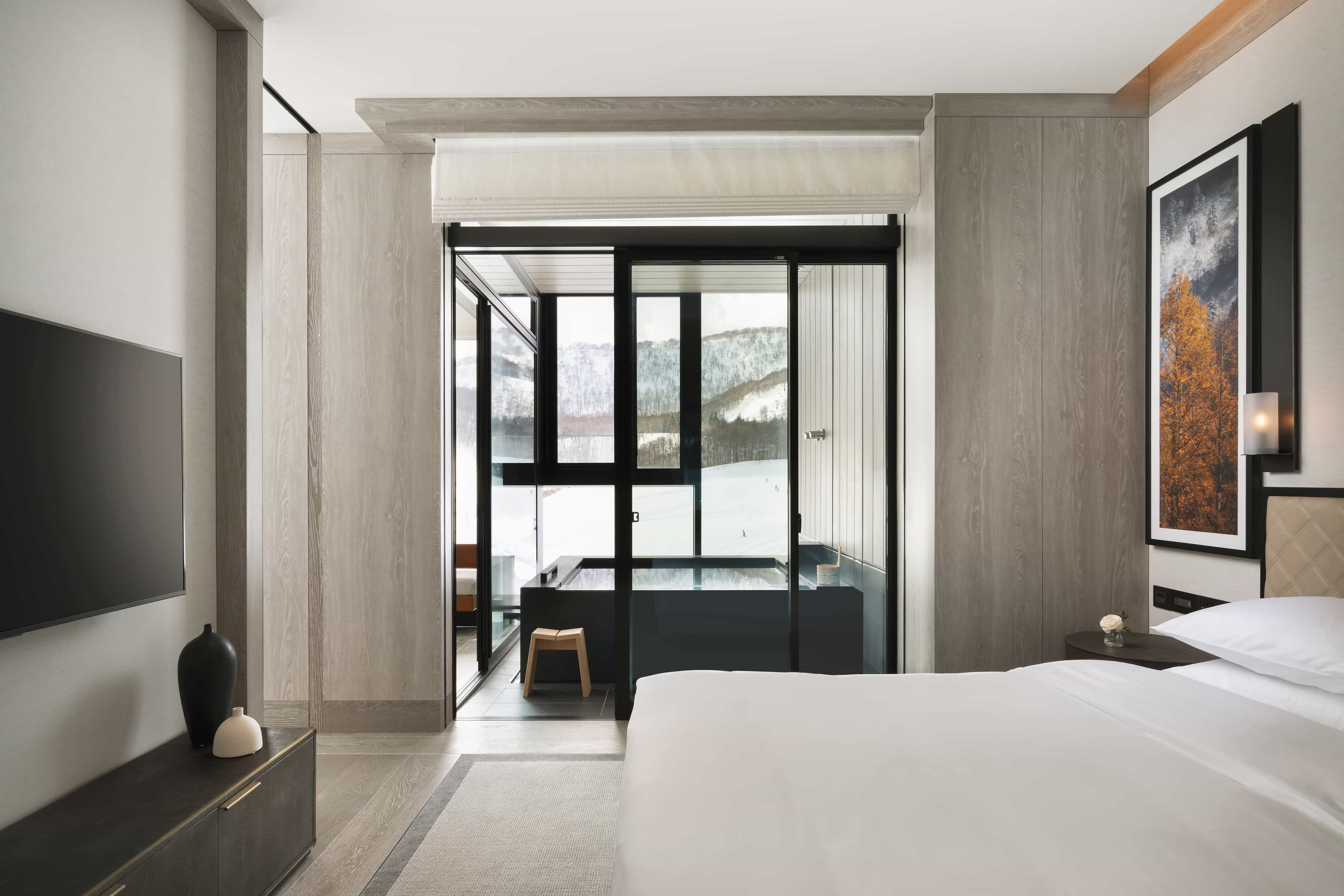 Sleek, minimalist hotel bedroom with a king bed, warm wood paneling, and a glass-enclosed soaking tub overlooking snow-covered mountains.