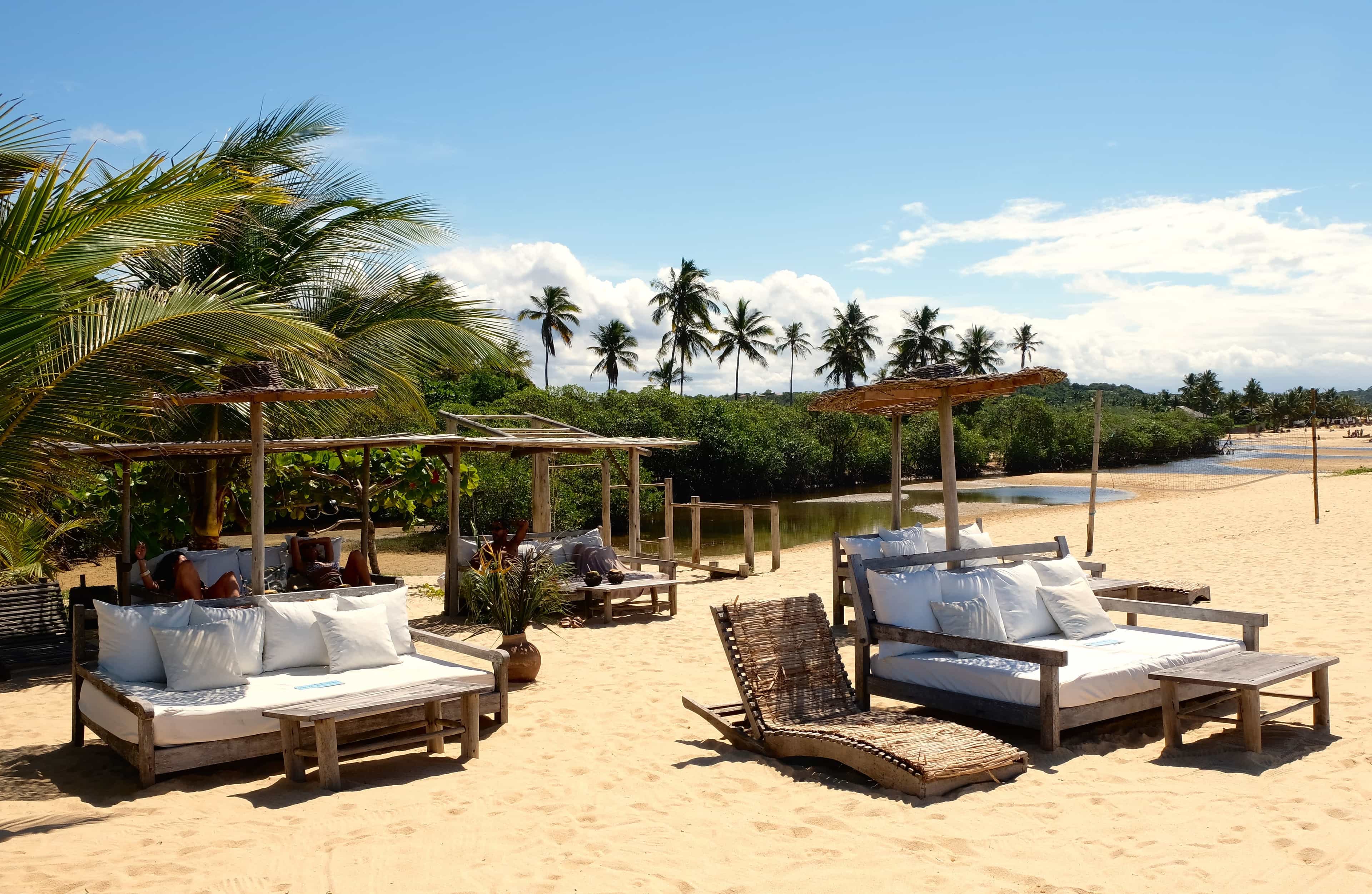 Rustic beach loungers with white cushions under wooden shade structures, set on golden sand with palm trees and a calm lagoon in the background under a bright blue sky.
