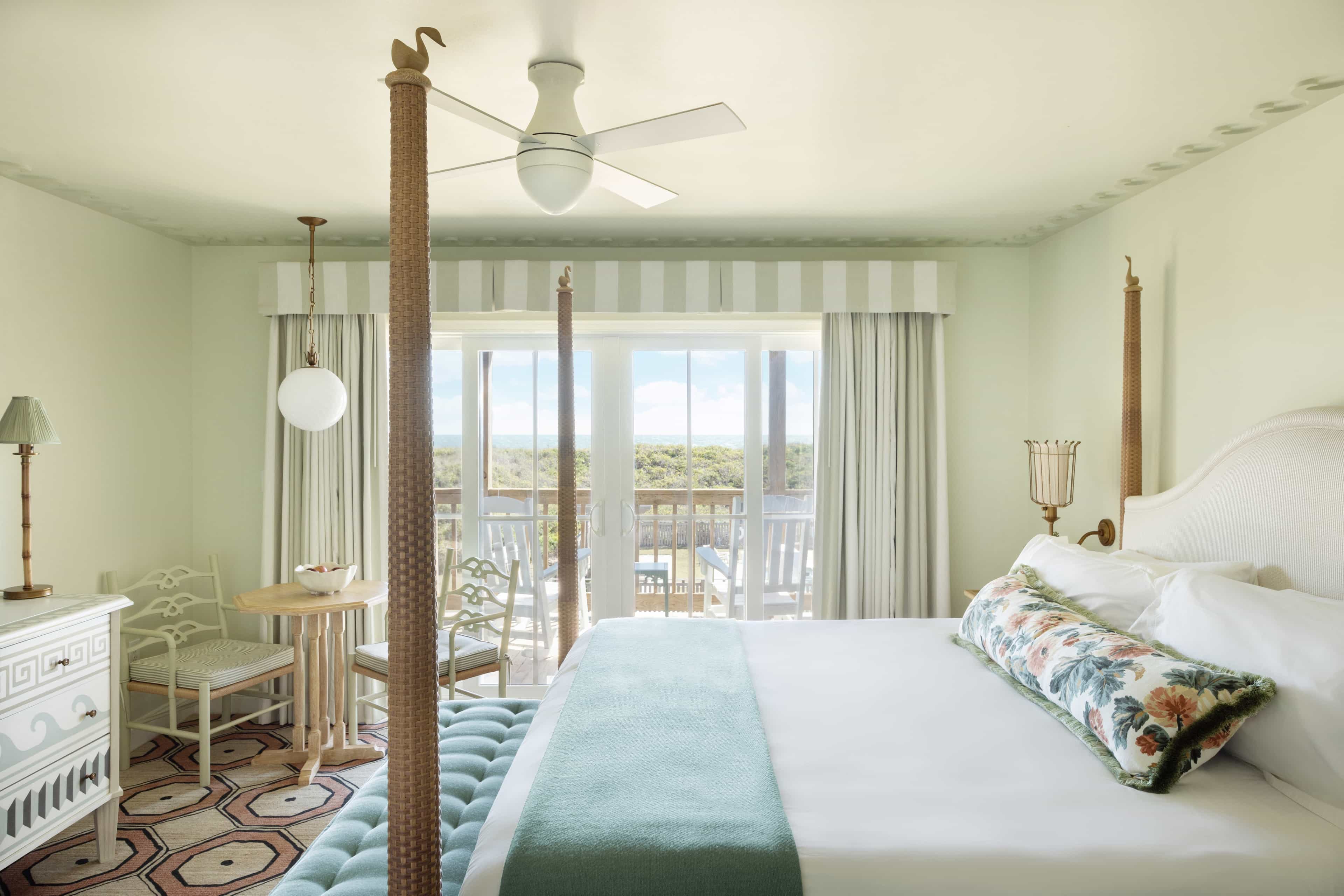 Bright coastal-style bedroom with a four-poster bed, soft green accents, patterned rug, and French doors opening onto a balcony with treetop and ocean views.