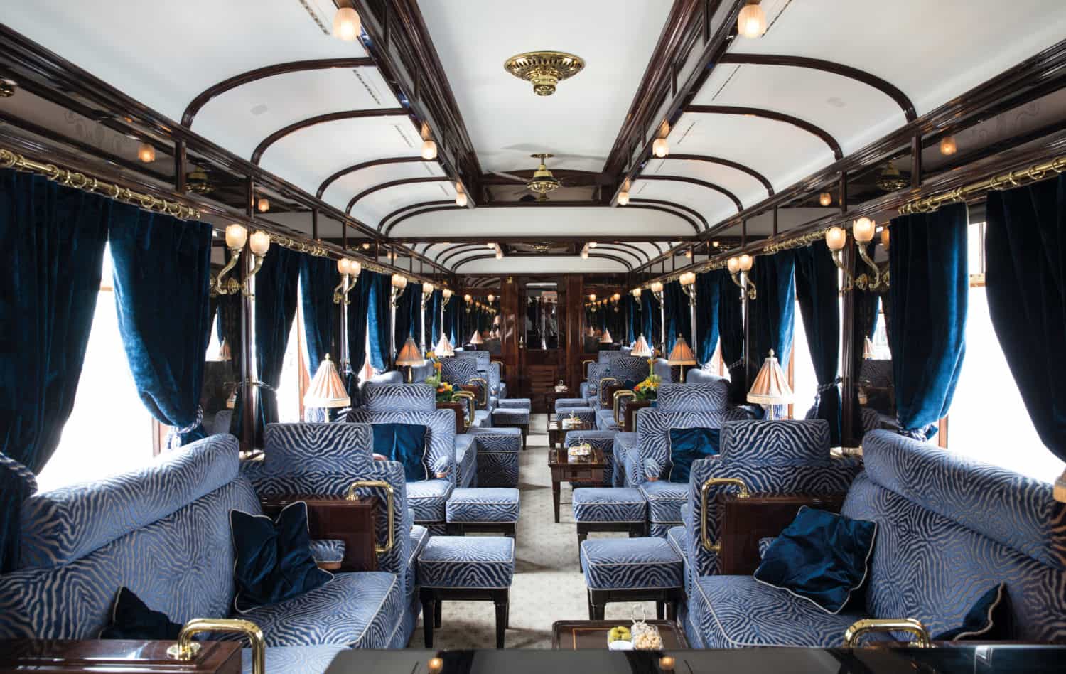 Elegant interior of the Venice Simplon-Orient-Express piano bar, with plush blue patterned armchairs, dark wood paneling, brass details, soft lamps, and arched ceilings creating a classic Art Deco-style train lounge.