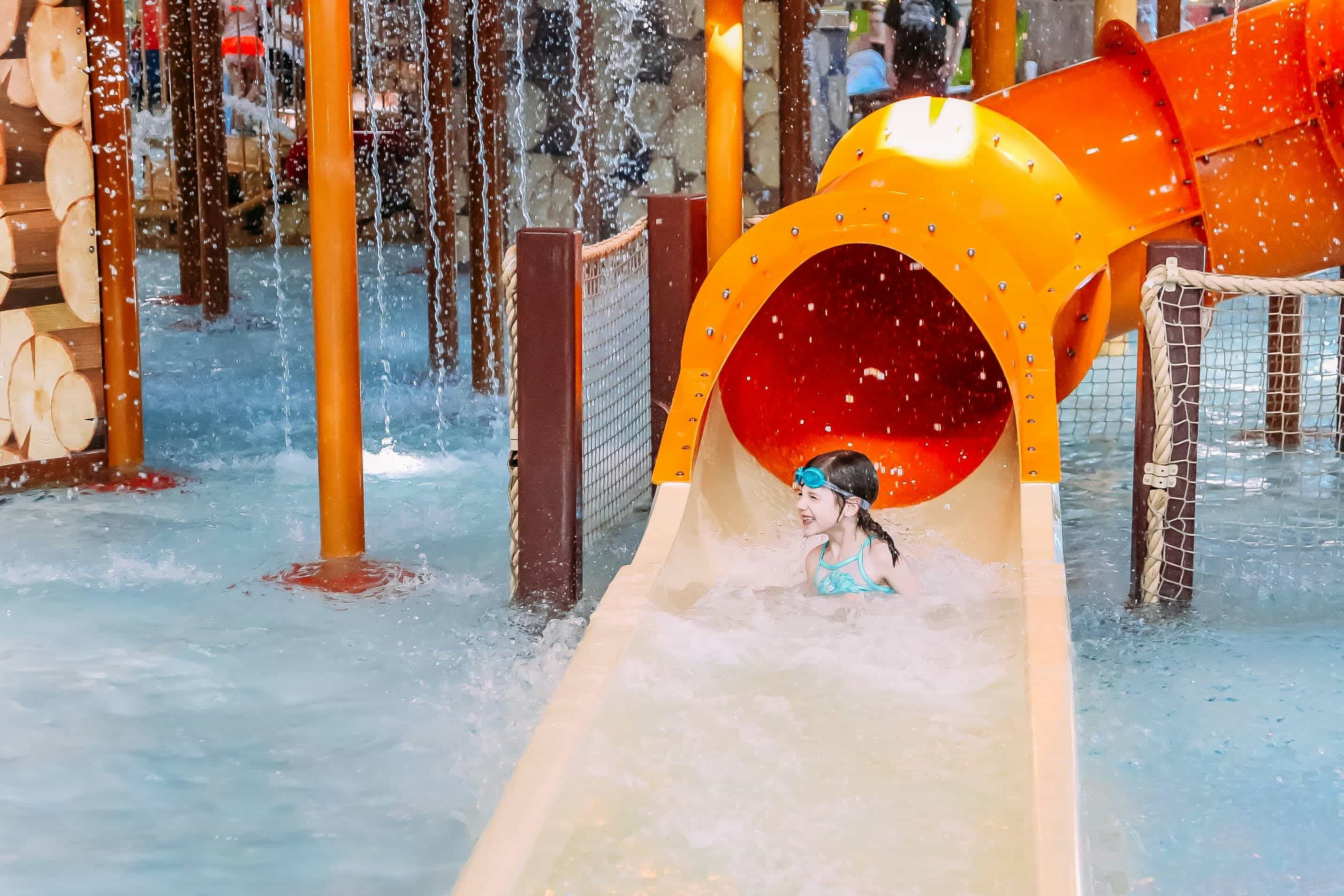 Young child smiling while coming down a bright orange water slide into a shallow pool.