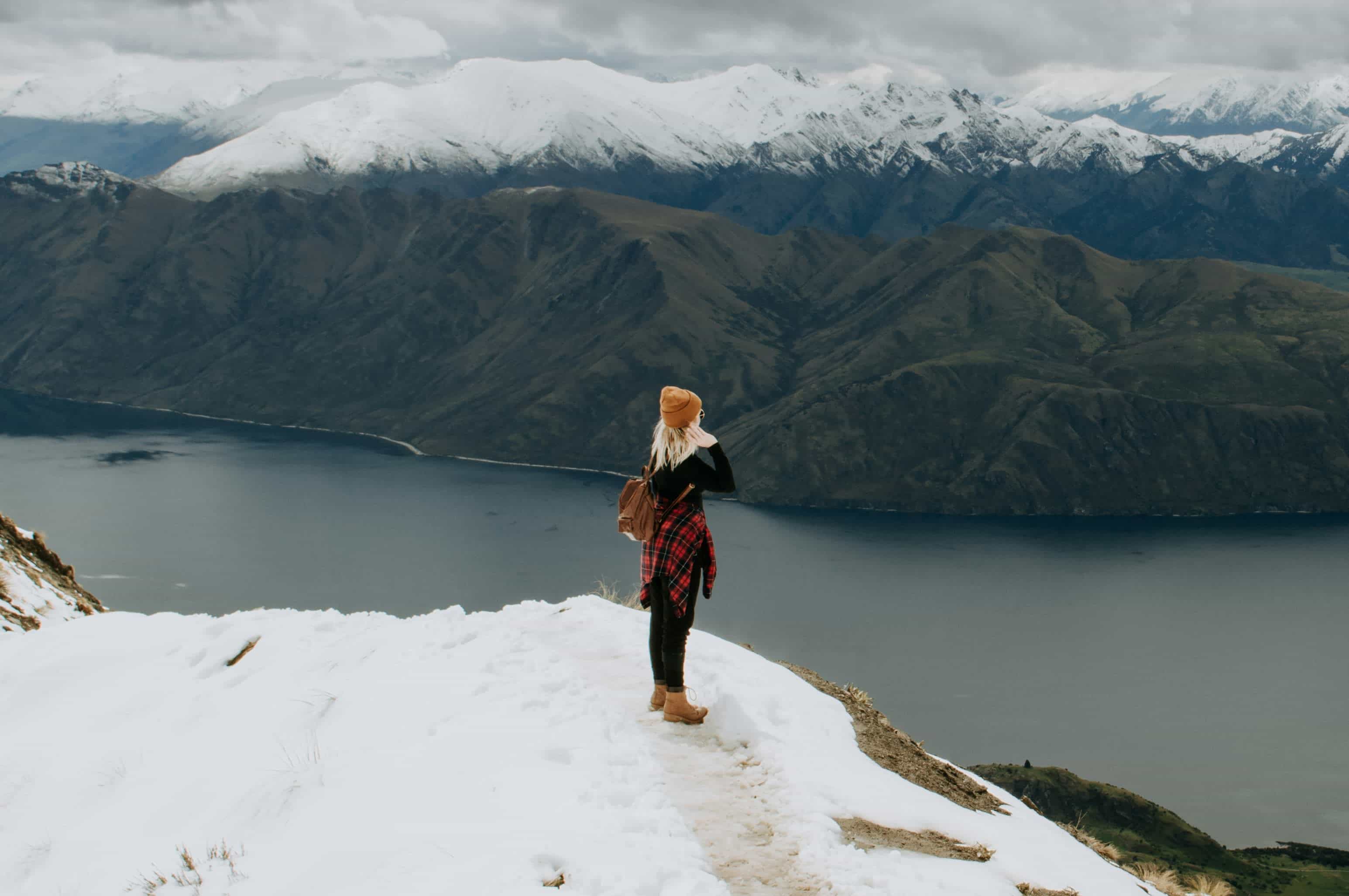 Person standing on a snowy cliff edge, looking out over a deep blue lake and dramatic snow-capped mountains under a cloudy sky.