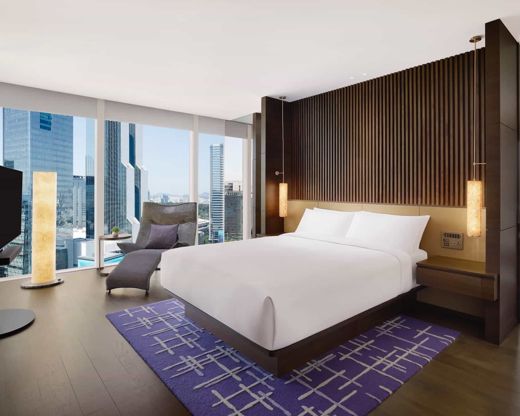 white bed up against a dark brown wood paneled wall with grey chair next to floor-to-ceiling windows