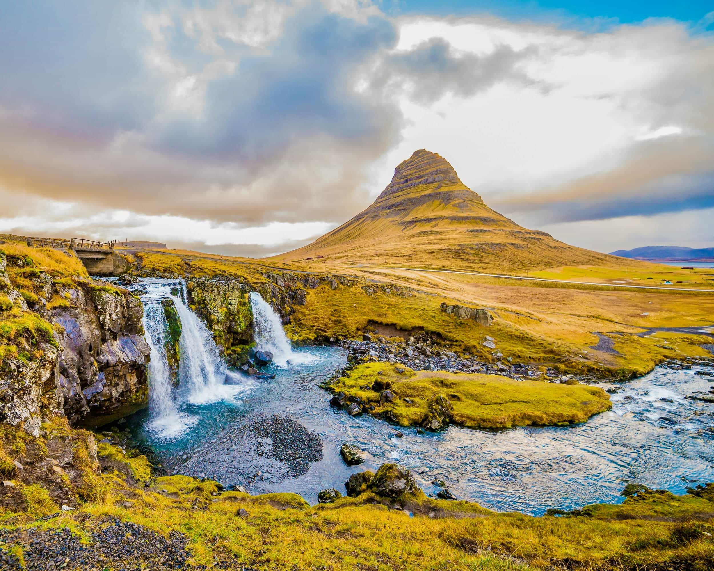 waterfalls next to a tall yellowish green mountain during day with a cloudy sky