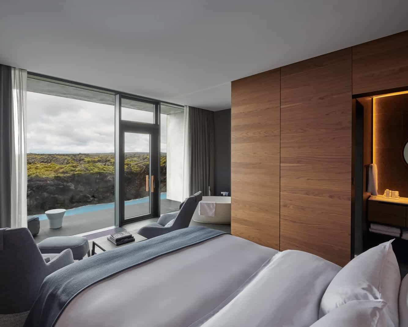 Hotel room with wood walls, white bed facing floor-to-ceiling windows, with two chairs and bathtub