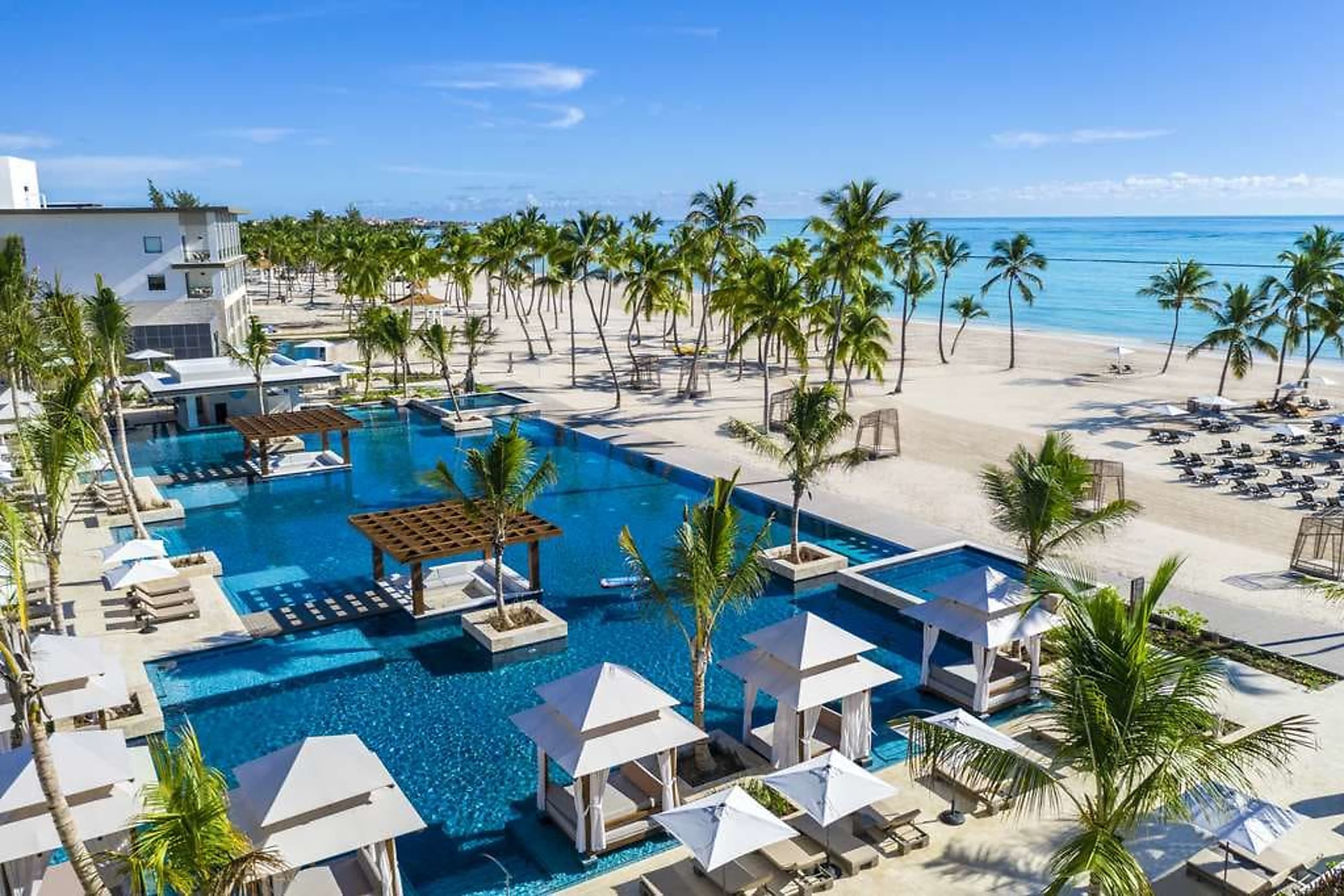 Expansive resort grounds with a large, palm tree-lined pool area blending into a white-sand beach
