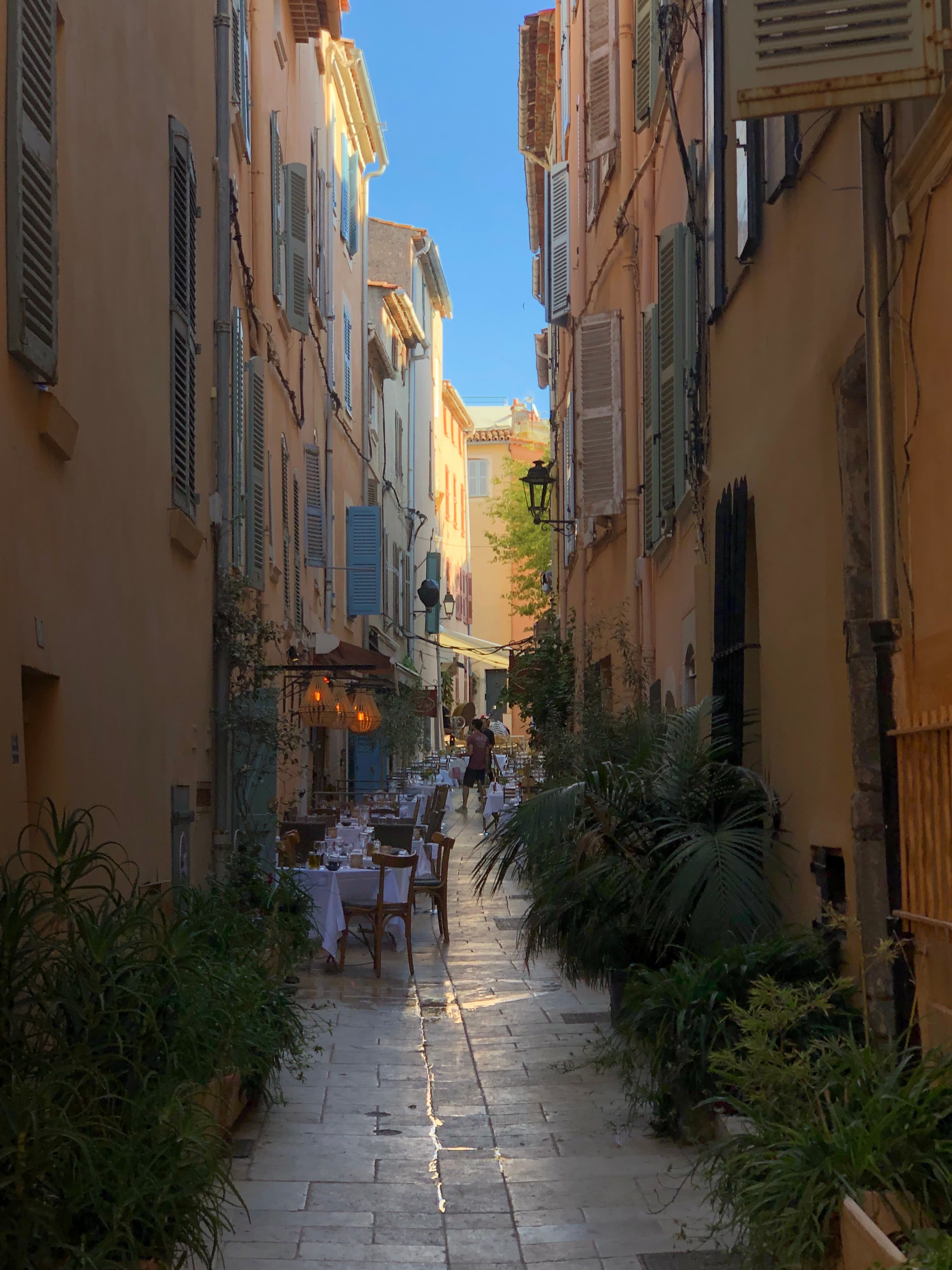 An alley way at dusk with outside tables, foliage and beige colored walls with the sun in the distance.