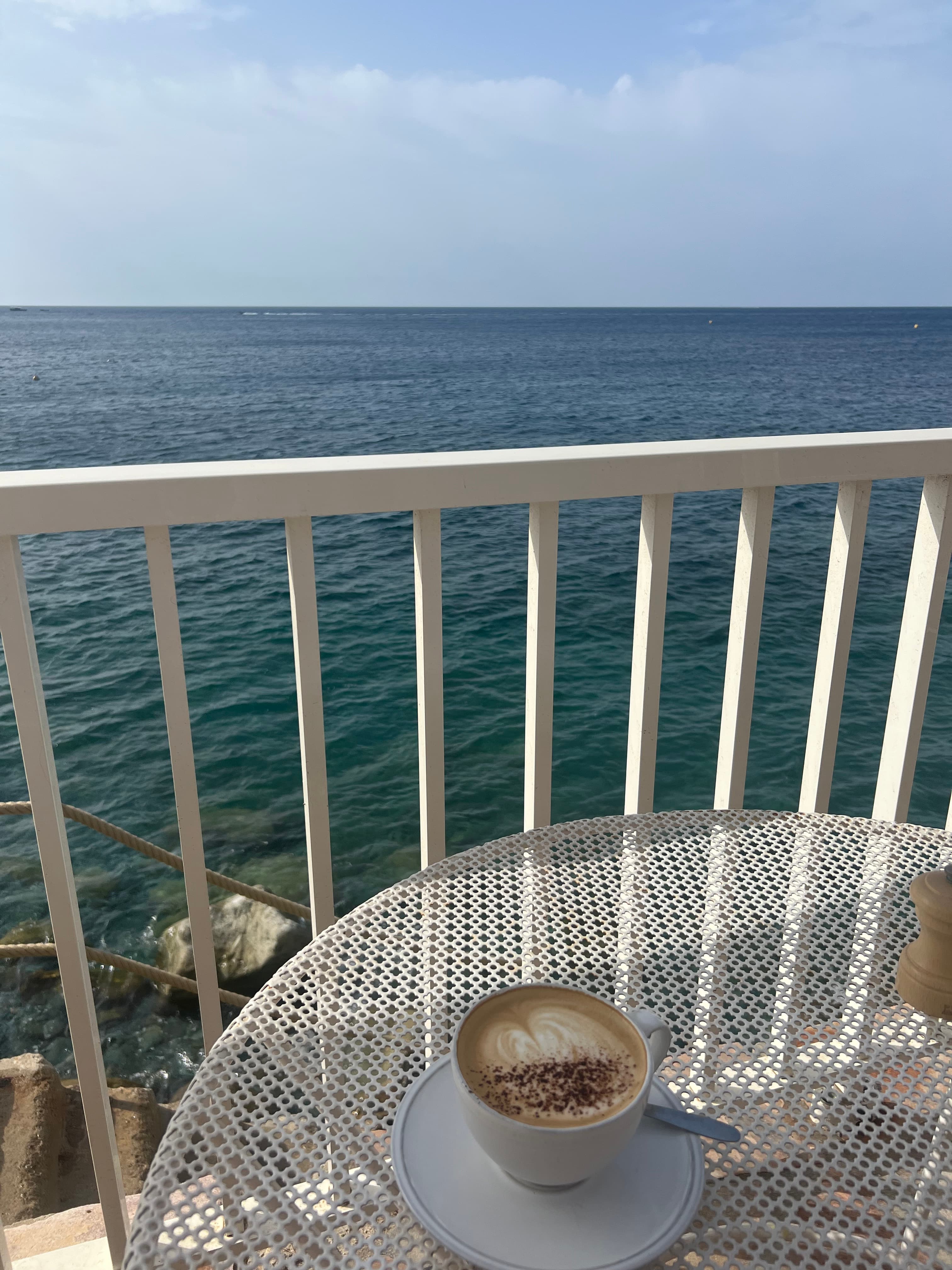An outdoor table on balcony with a coffee and a view of the ocean in the distance.