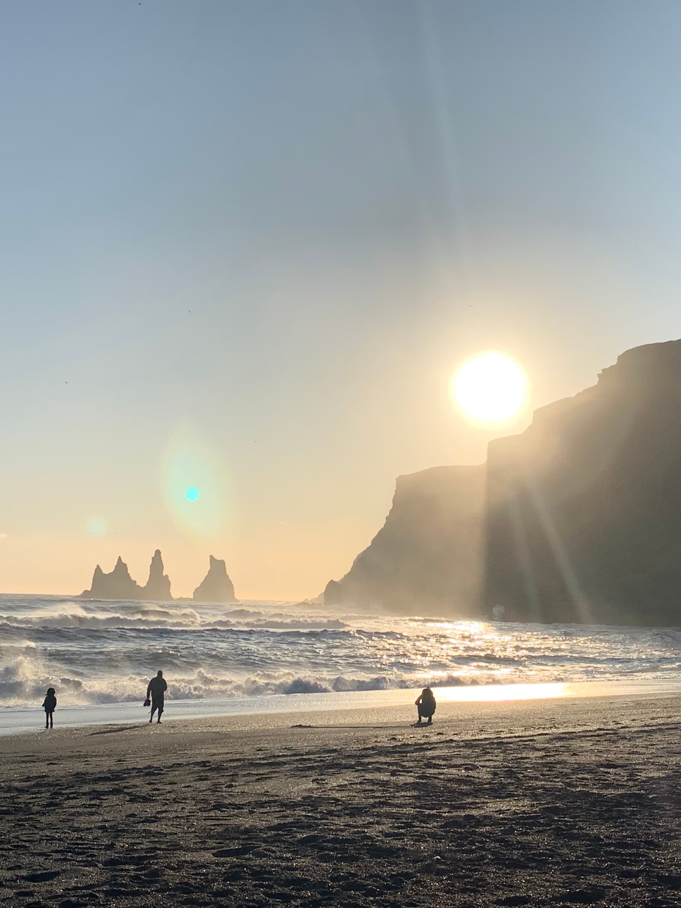 People on a beach at sunset with the ocean and a shoreline with cliffs in the distance.