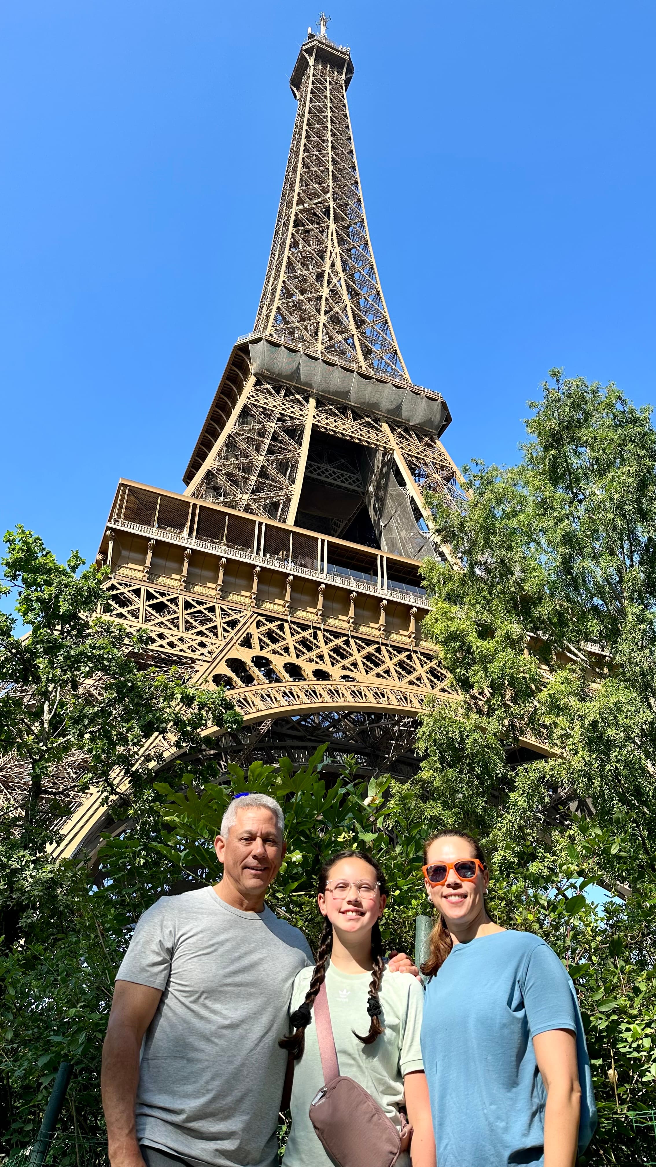 A family posing outside of the Eiffel Tower on a sunny day with foliage in the distance.