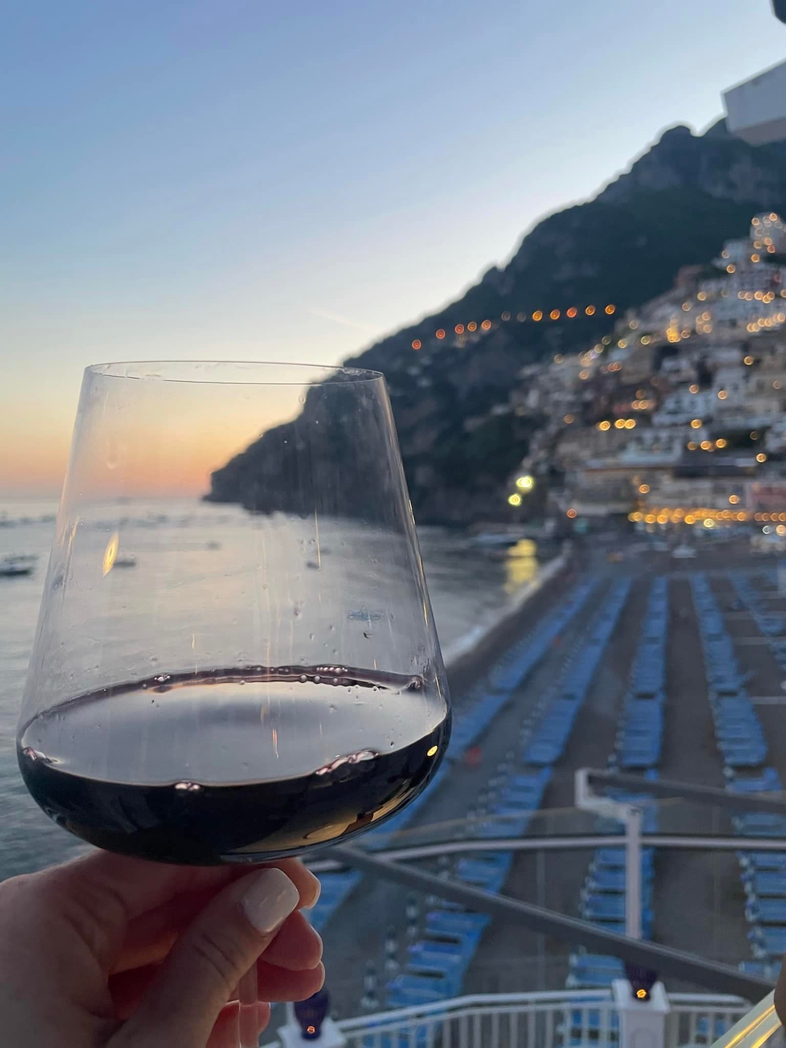 A close-up image of a hand holding a glass of red wine overlooking a beach and ocean at sunset
