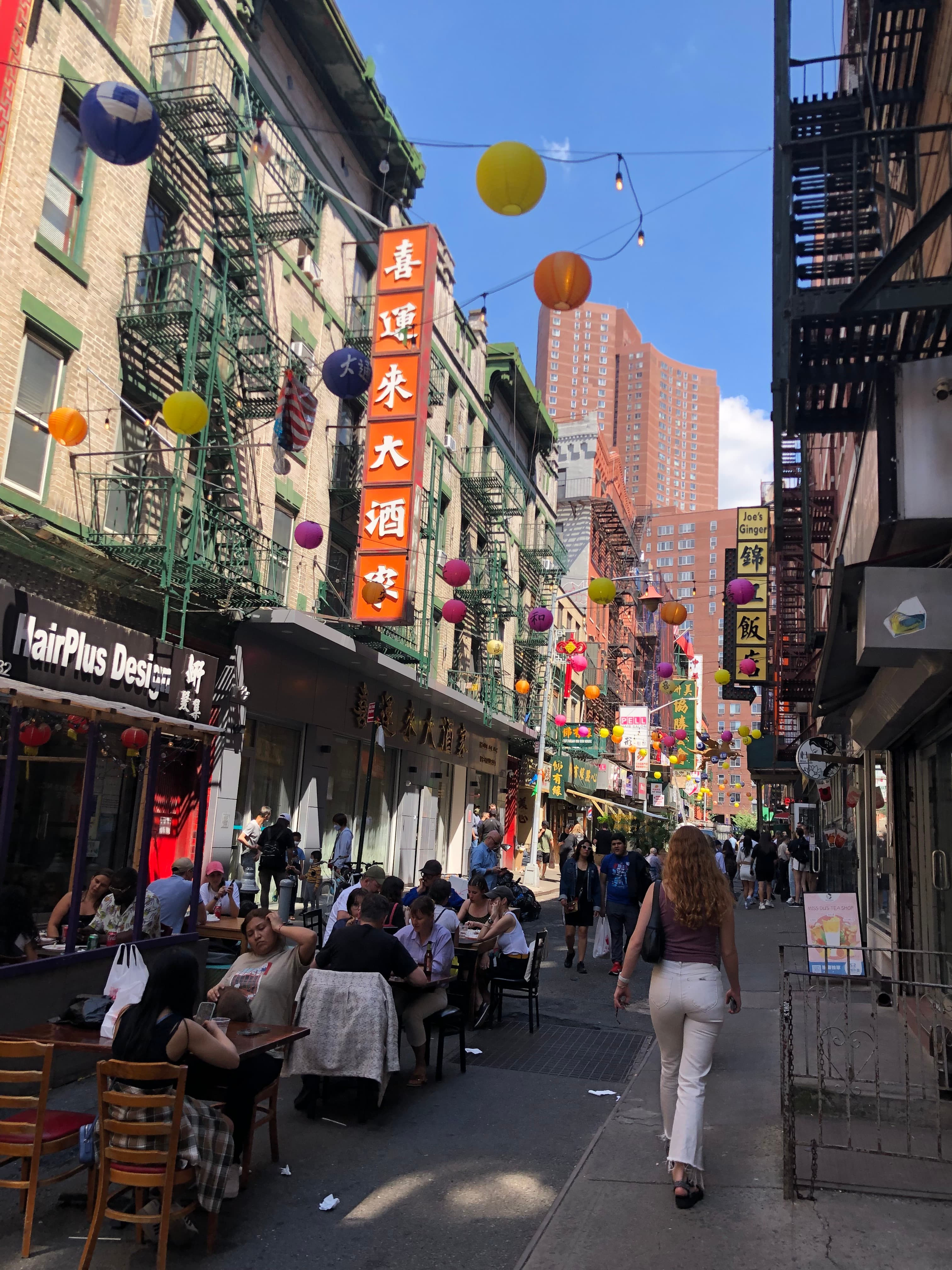 A street scene of a city's China town, with people sitting at outdoor cafés, hanging lanterns, shops and pedestrians on a sunny day.