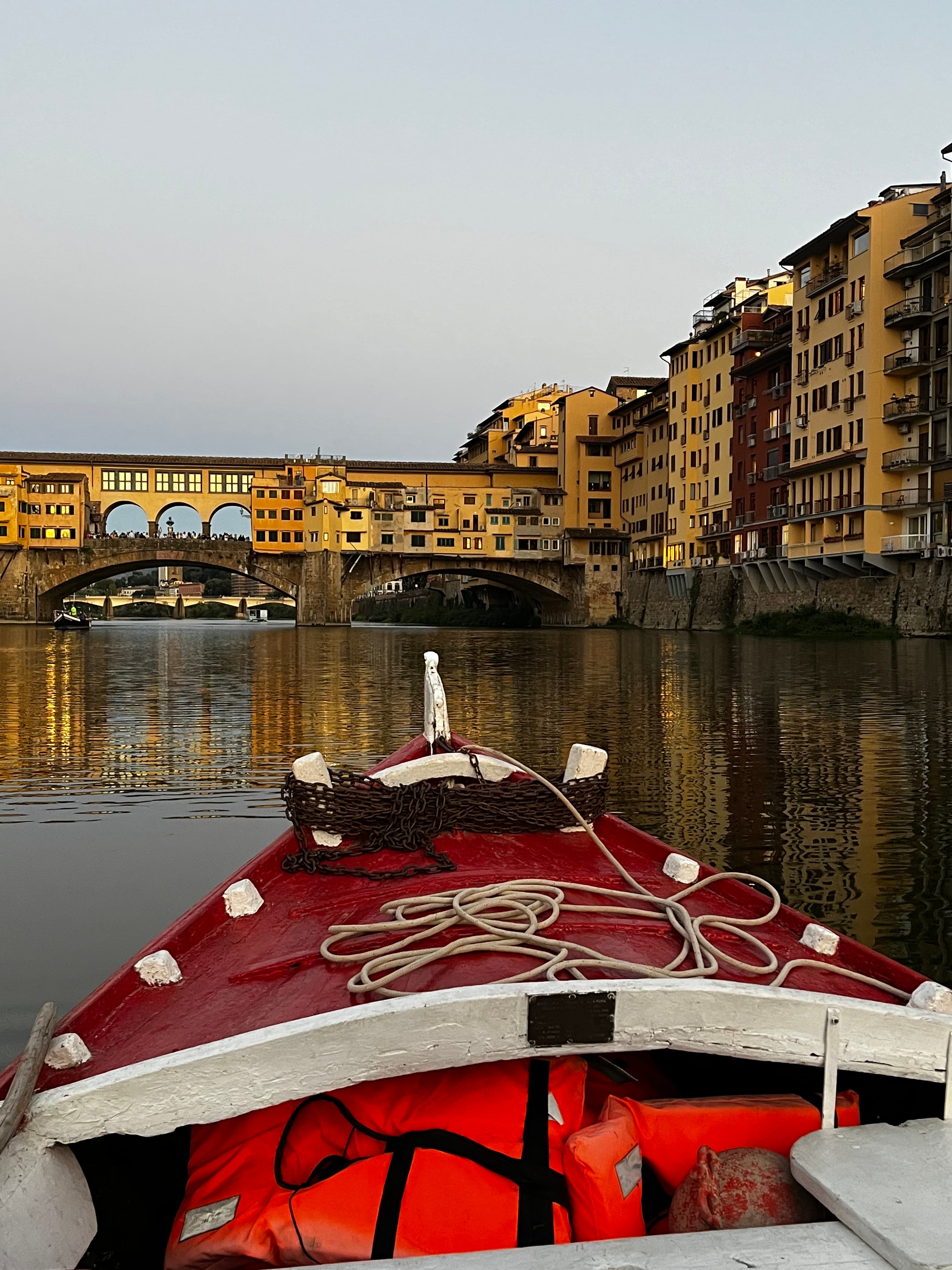 A point of view shot of the bow of a small, red boat on a river, with a bridge and city buildings in the distance.