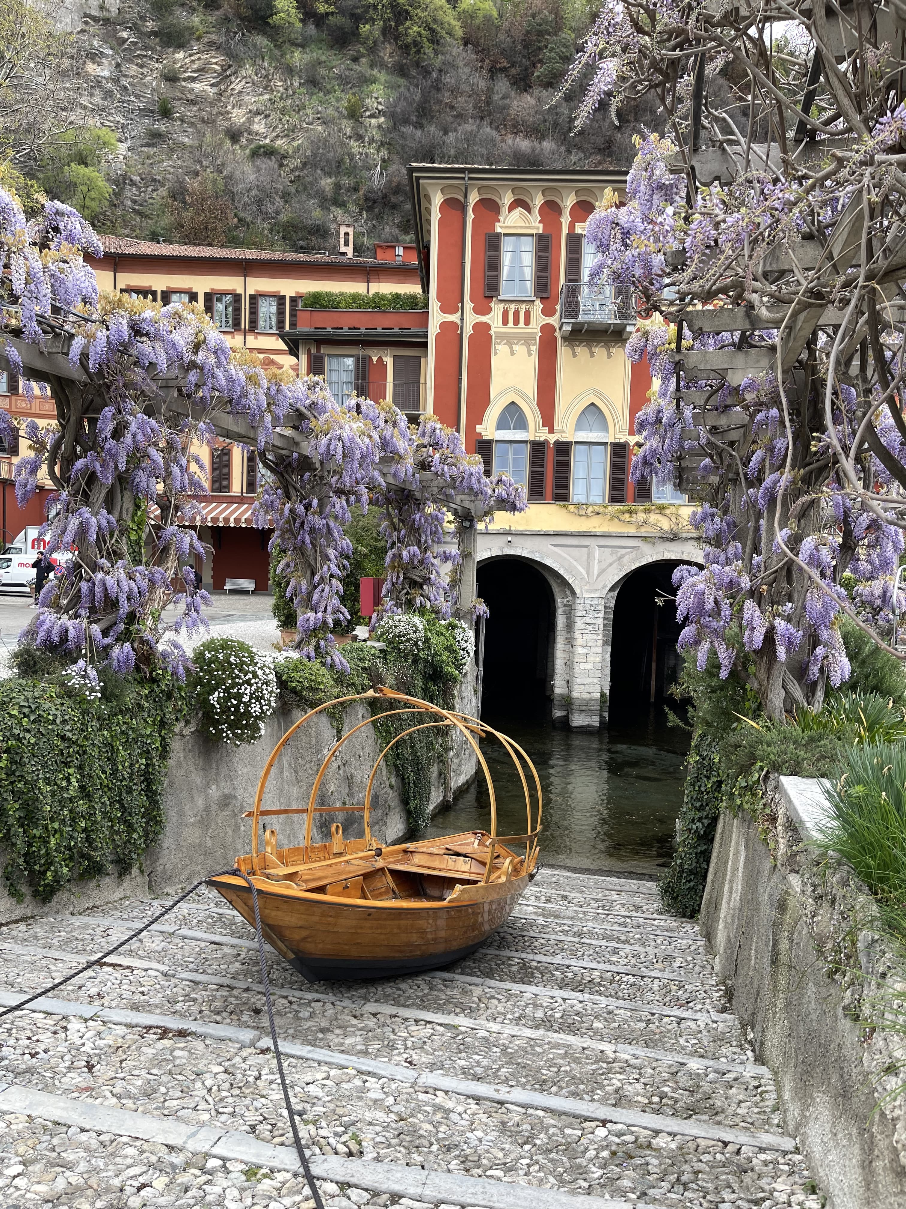 Boat on an angled stone deck next to a river framed with wisteria and a red-and-yellow building.