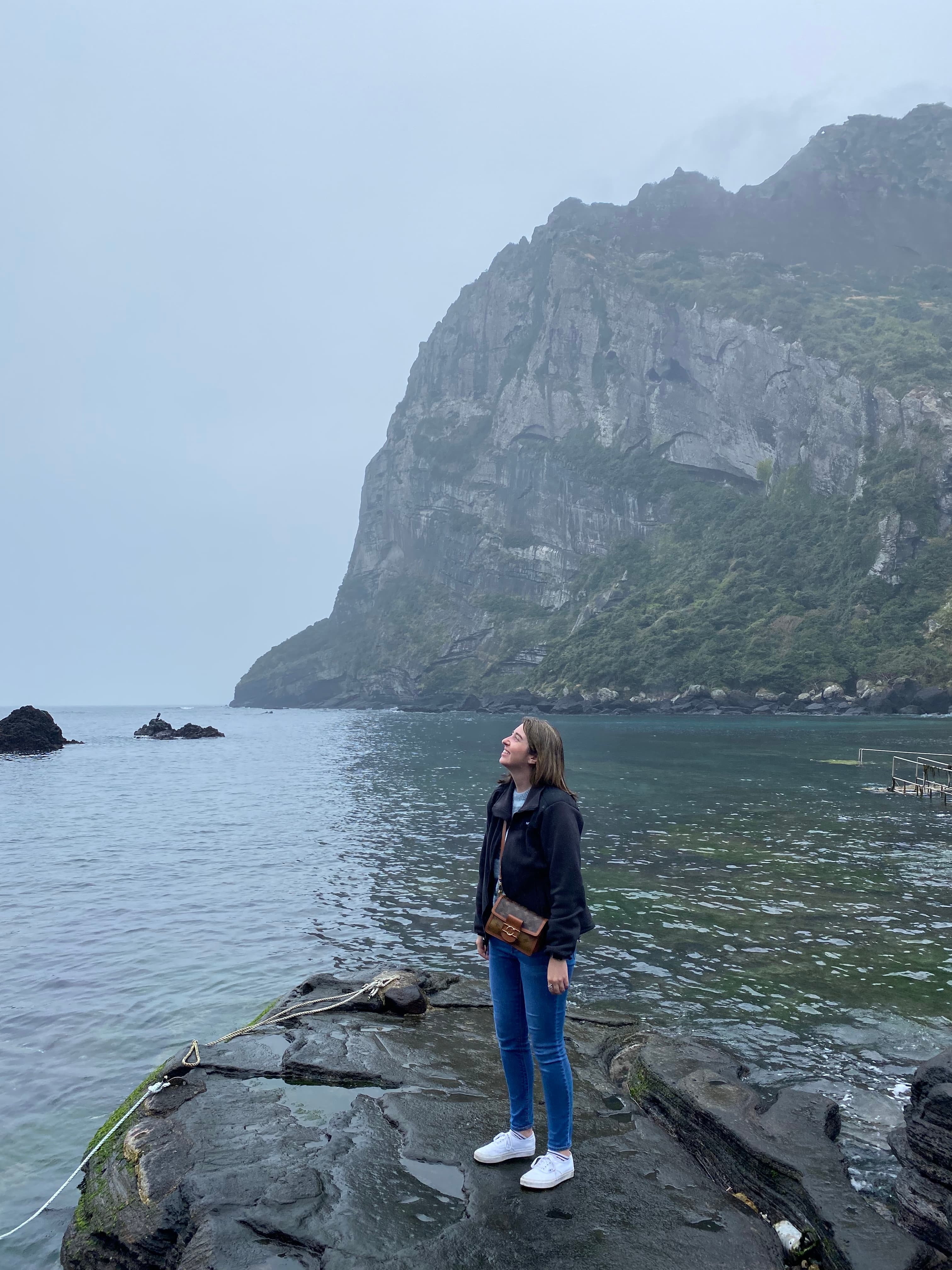Advisor posing for an image on a rock formation with the ocean in the distance on a cloudy day.