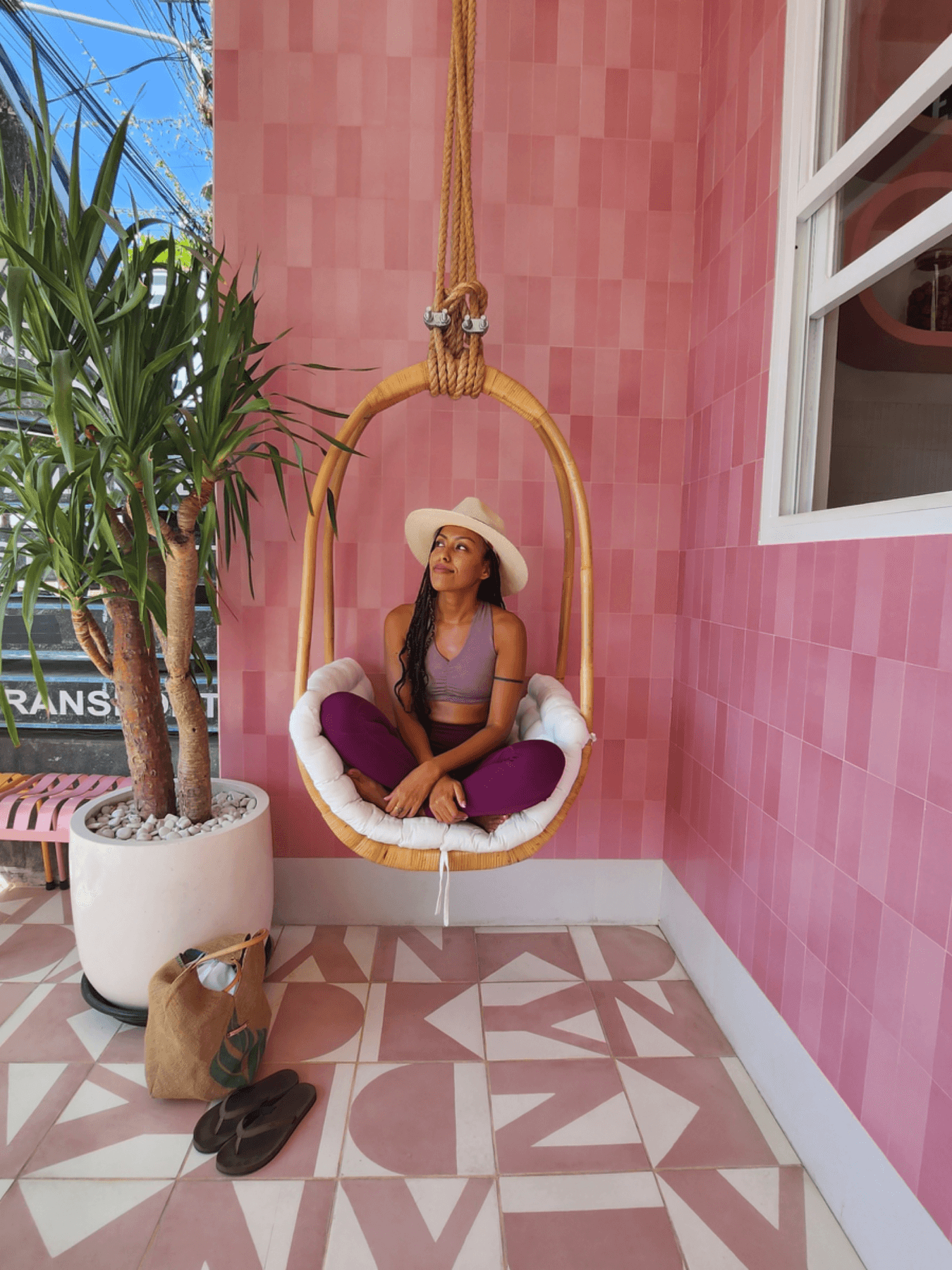 Suzan, sitting cross legged in a hanging chair made of bamboo in front of a pink, patterned wall.