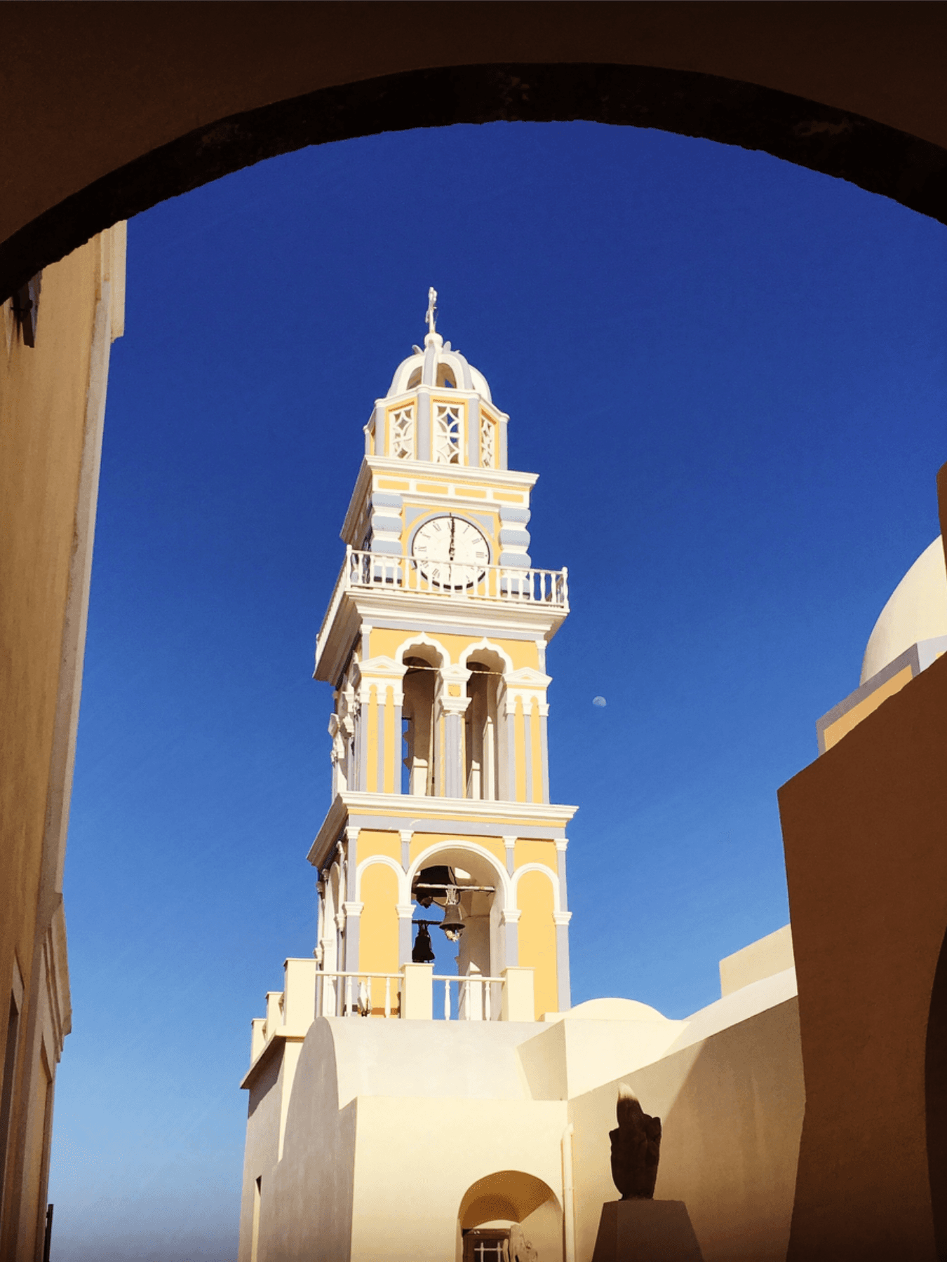 A photo of a tan and white church bell tower from beneath an archway on a sunny day.