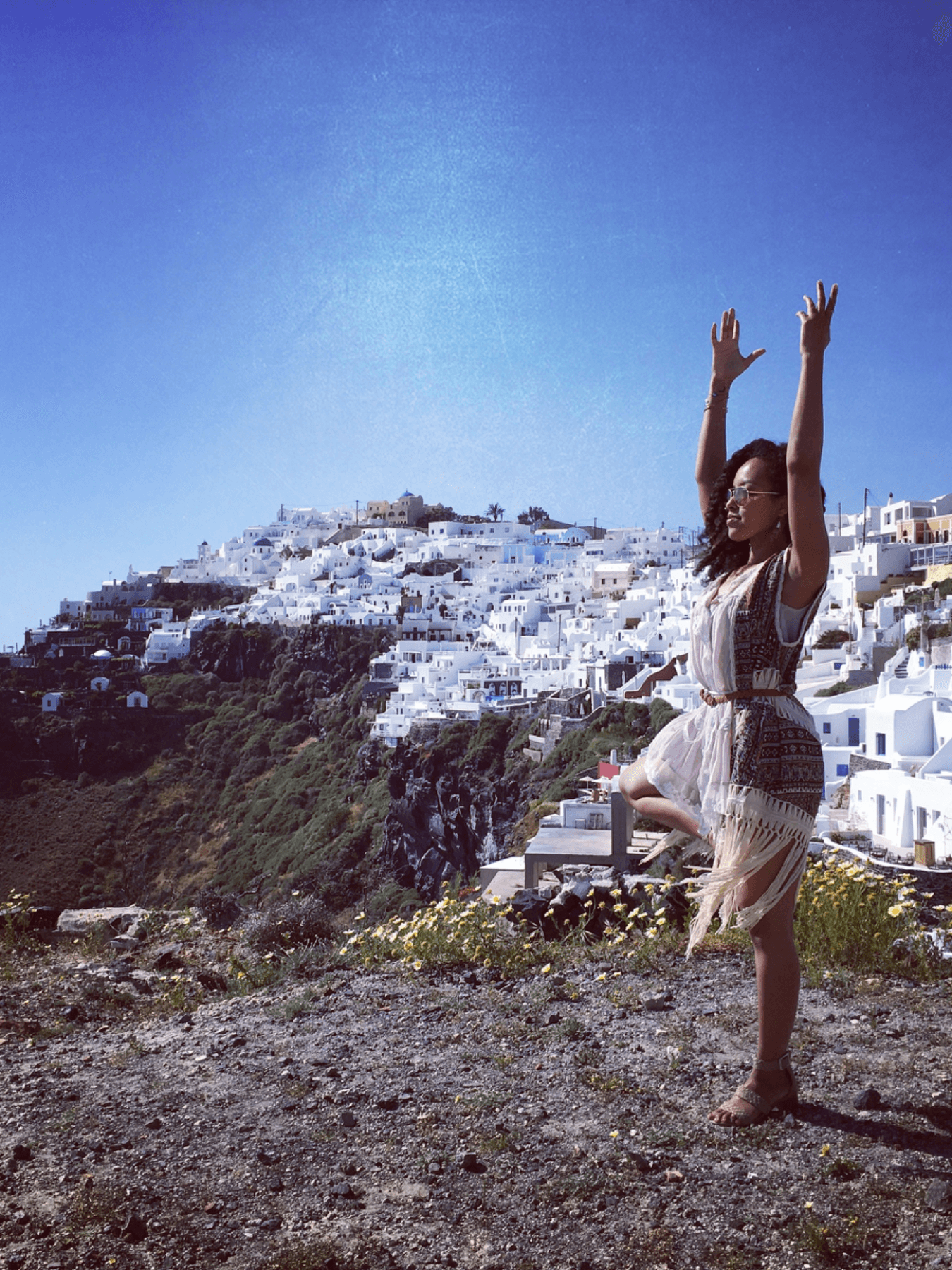 Suzan doing a yoga pose on a cliff overlooking a Greek town consisting of white clay houses.