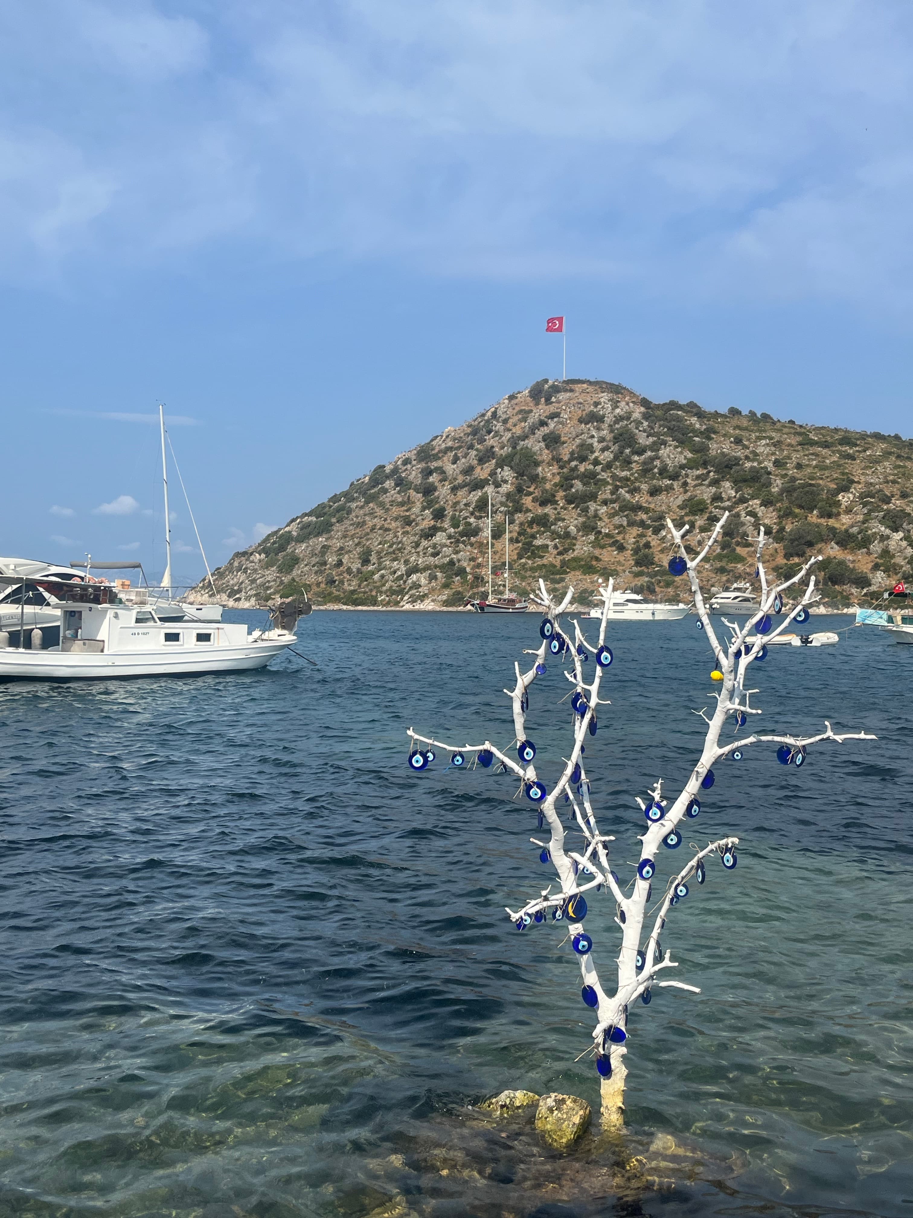 Image of a small white tree decorated with blue ornaments coming out of the ocean with white boats floating in the distance