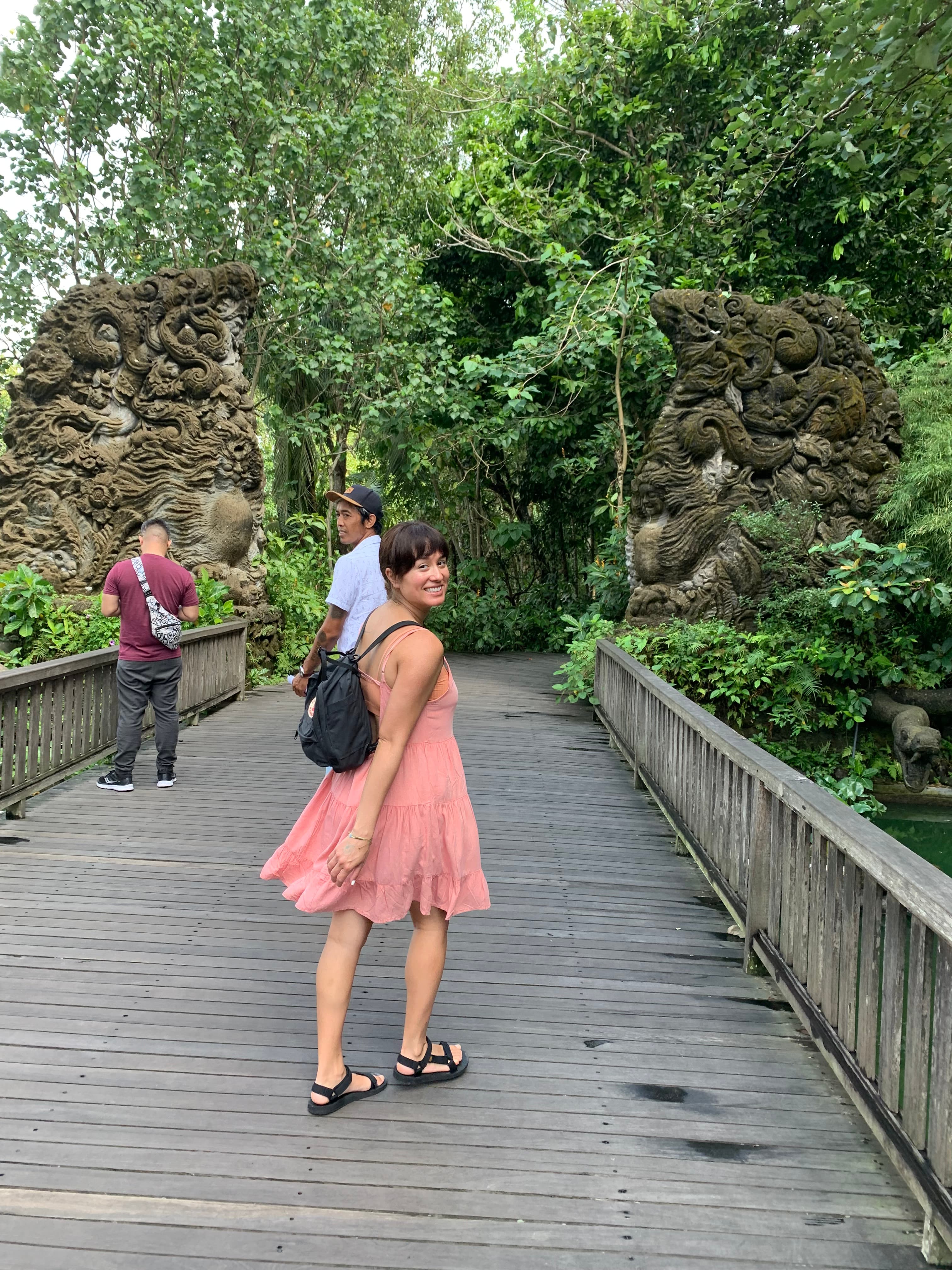 Michelle in a pink dress carrying a black backpack and smiling for a photo on a wide wooden bridge surrounded by trees