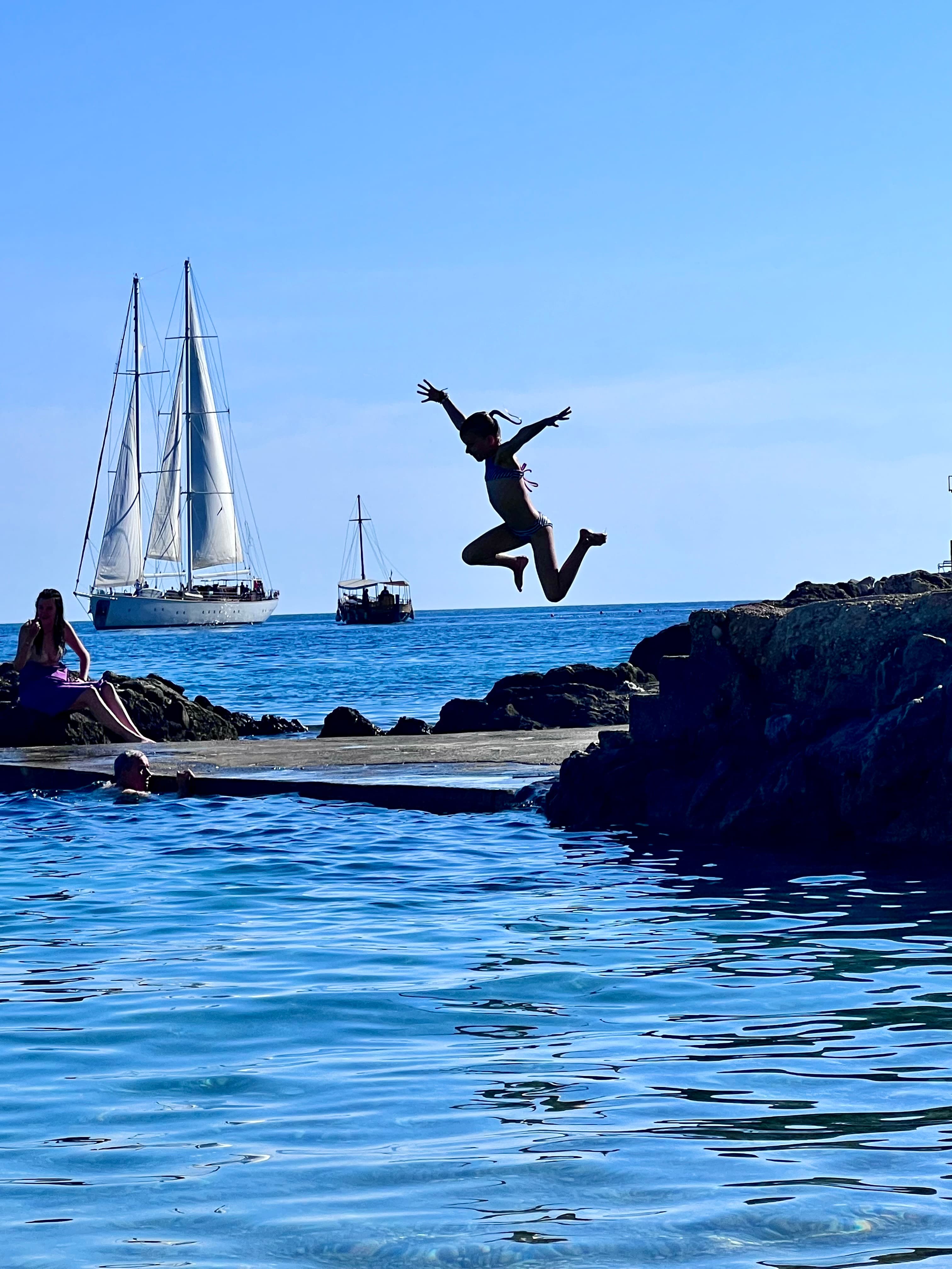 Image of Elizabeth jumping from rocks into the sea with a large sailboat visible on the horizon