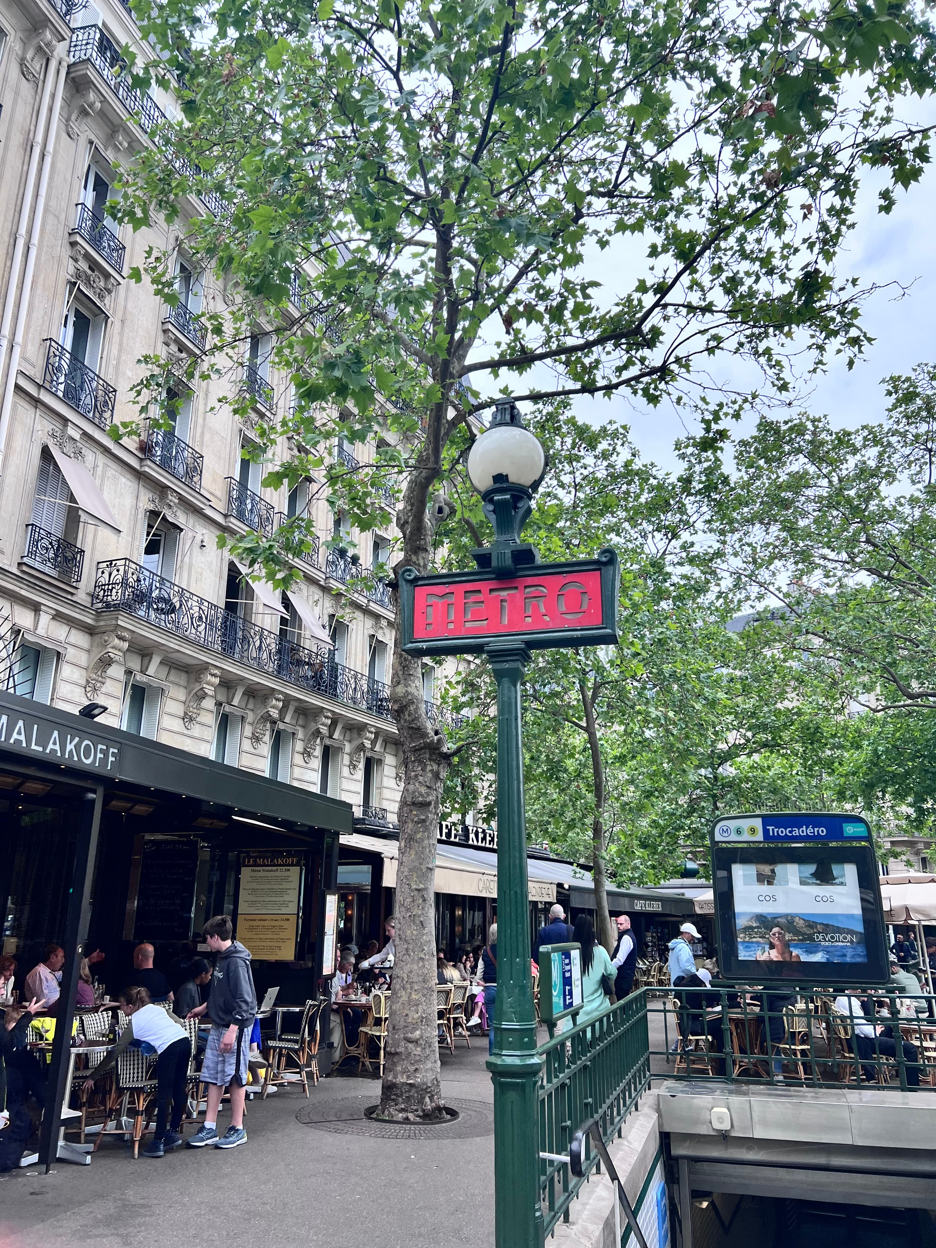 View of a Paris metro station sign on the street with trees and a restaurant patio behind it