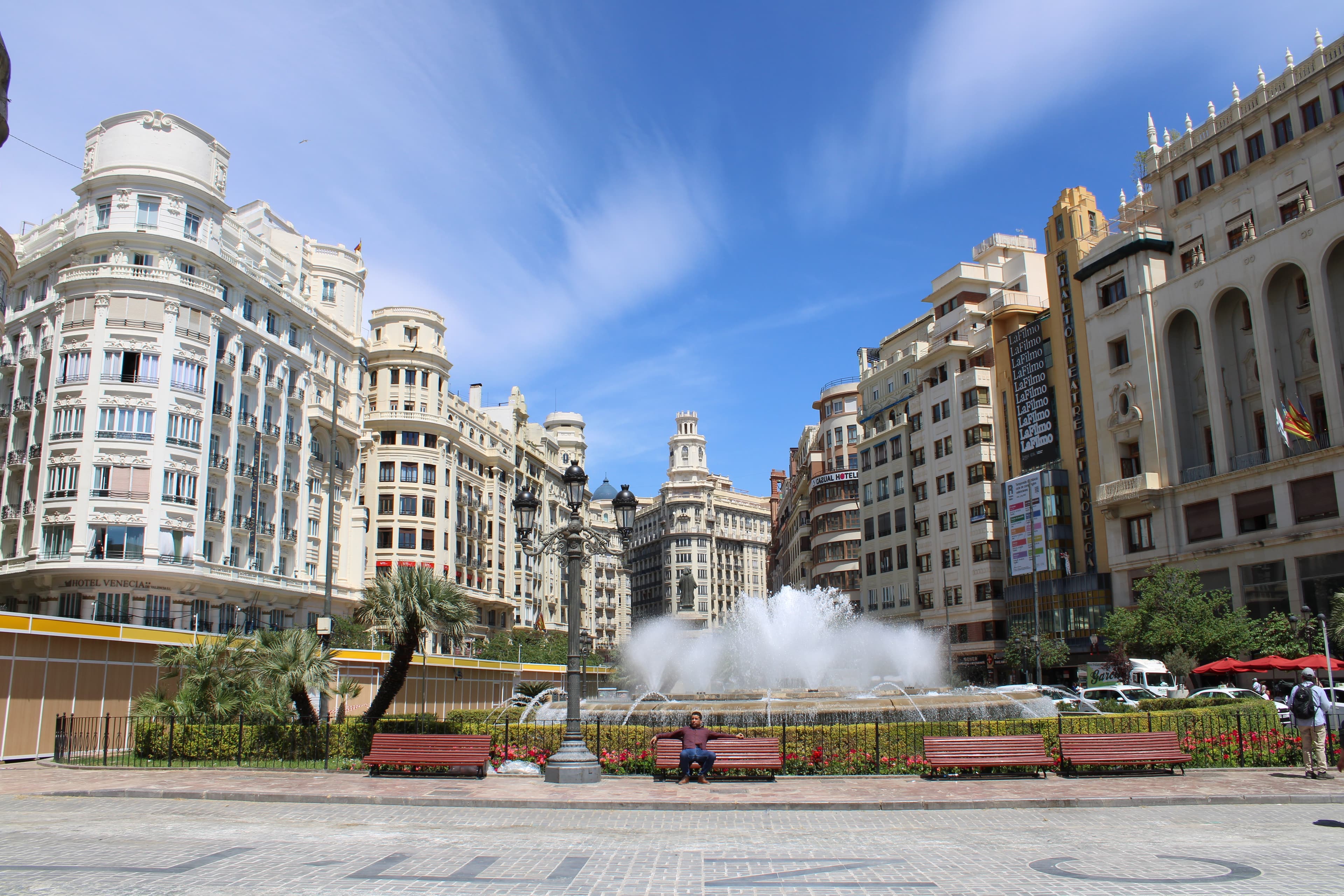 An image of a town square with a water fountain and classic buildings in the distance on a sunny day.