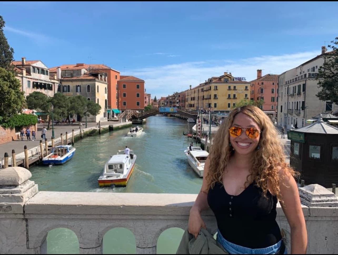 Maria in sunglasses and a black top posing on a bridge with colorful buildings and boats on a canal behind her